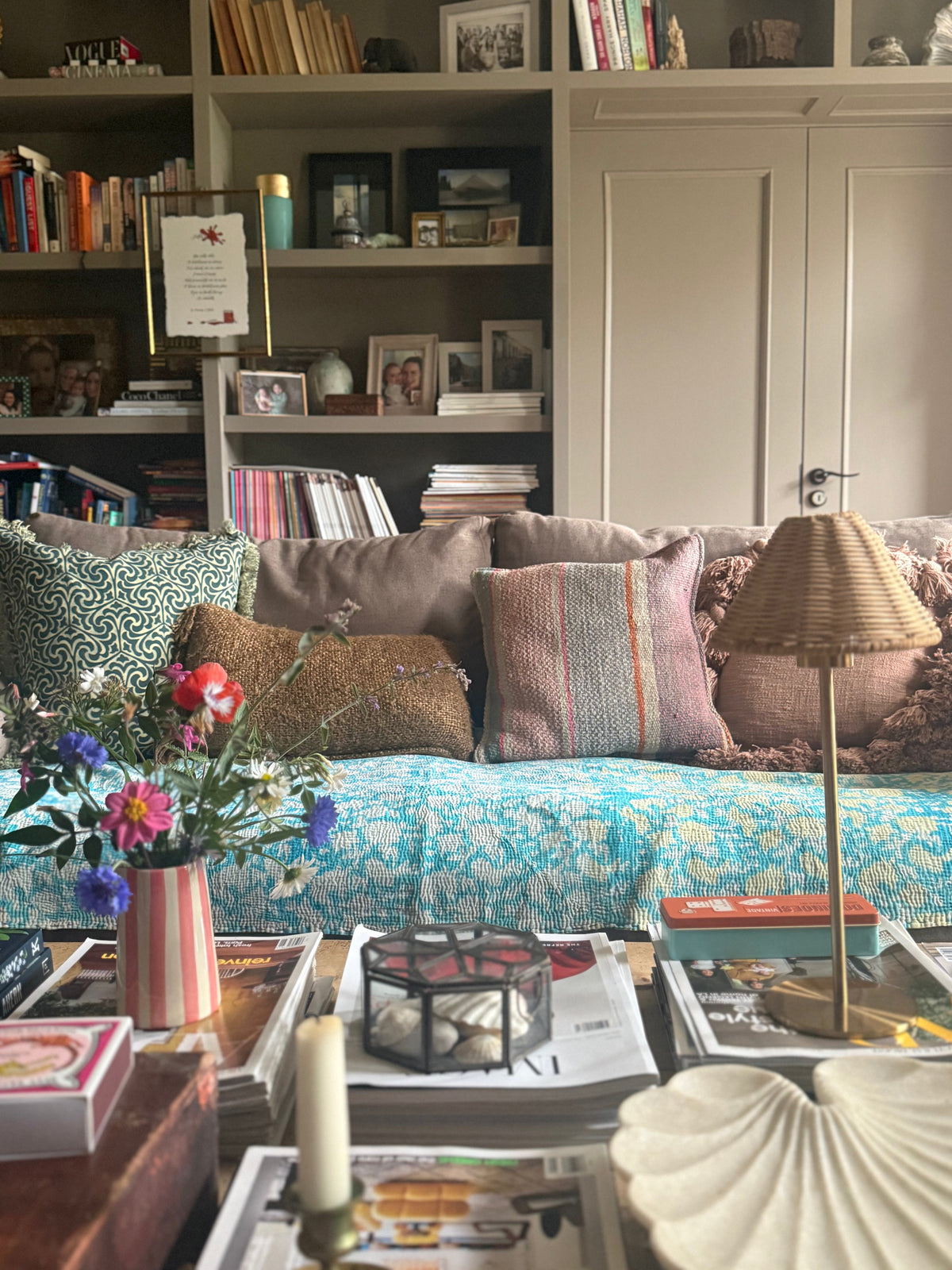 Eclectic living room with a sofa with mismatched cushions, colourful kantha throw, coffee table with books and decorative elements, a cordless rattan lamp on the table with bookshelves in the background filled with books and decor.