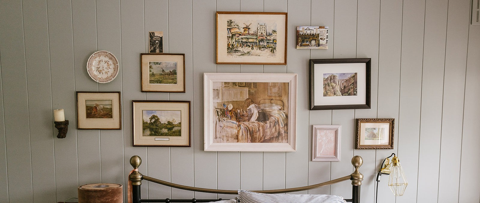 A tongue and grooved panelled light coloured wall with a gallery wall of vintage framed paintings, prints, vintage plates and wooden candleholder artfully arranged over a bed by Amber + Willow