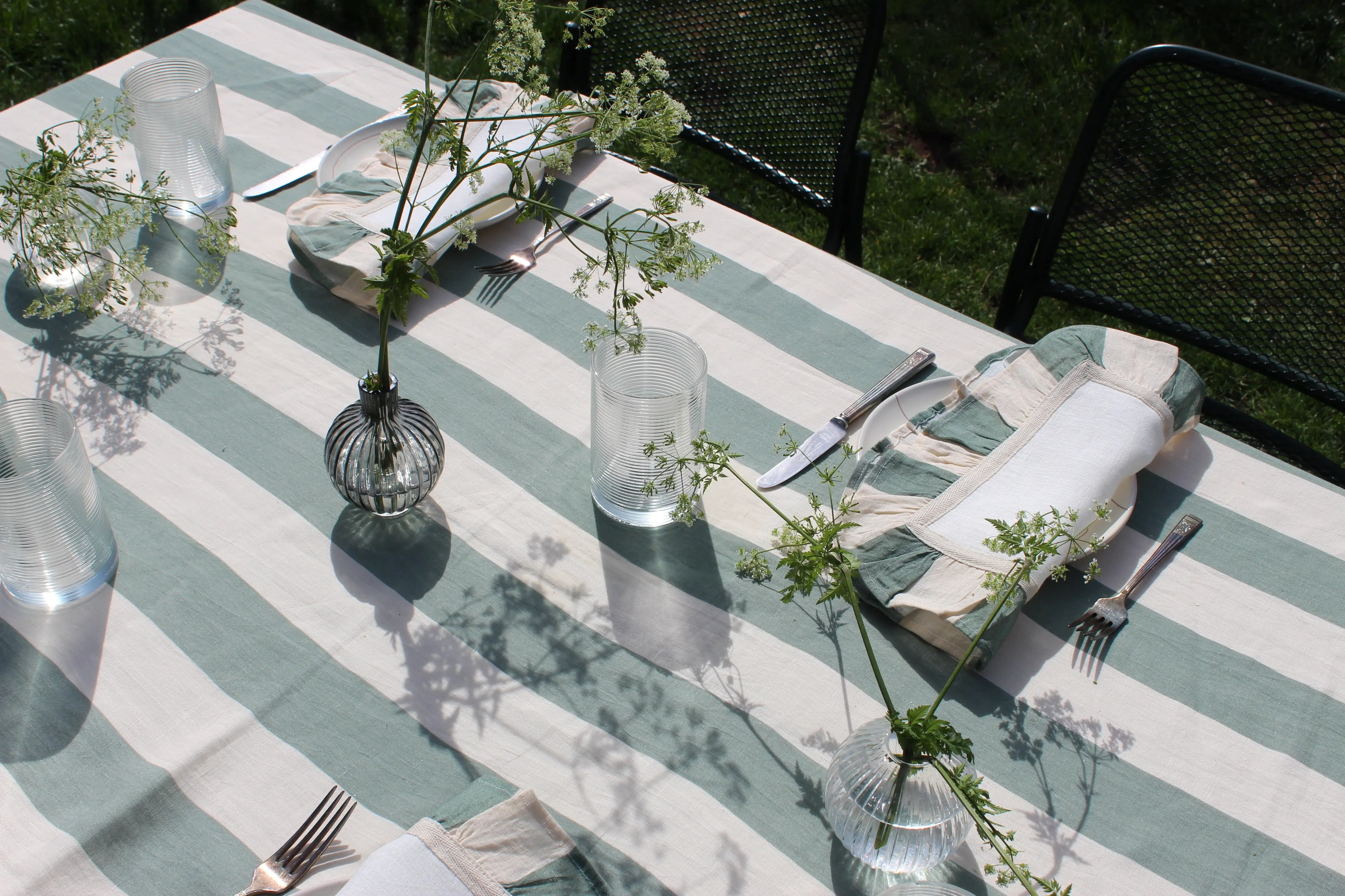 Sunny outdoor table setting with green and white striped table cloth,matching napkins, greenery in clear vases