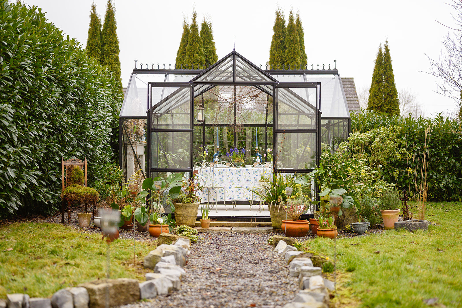Image of a charming black framed glass house in a country garden with curved gravel path leading up to it and greenery all around. Photo taken by Storybord