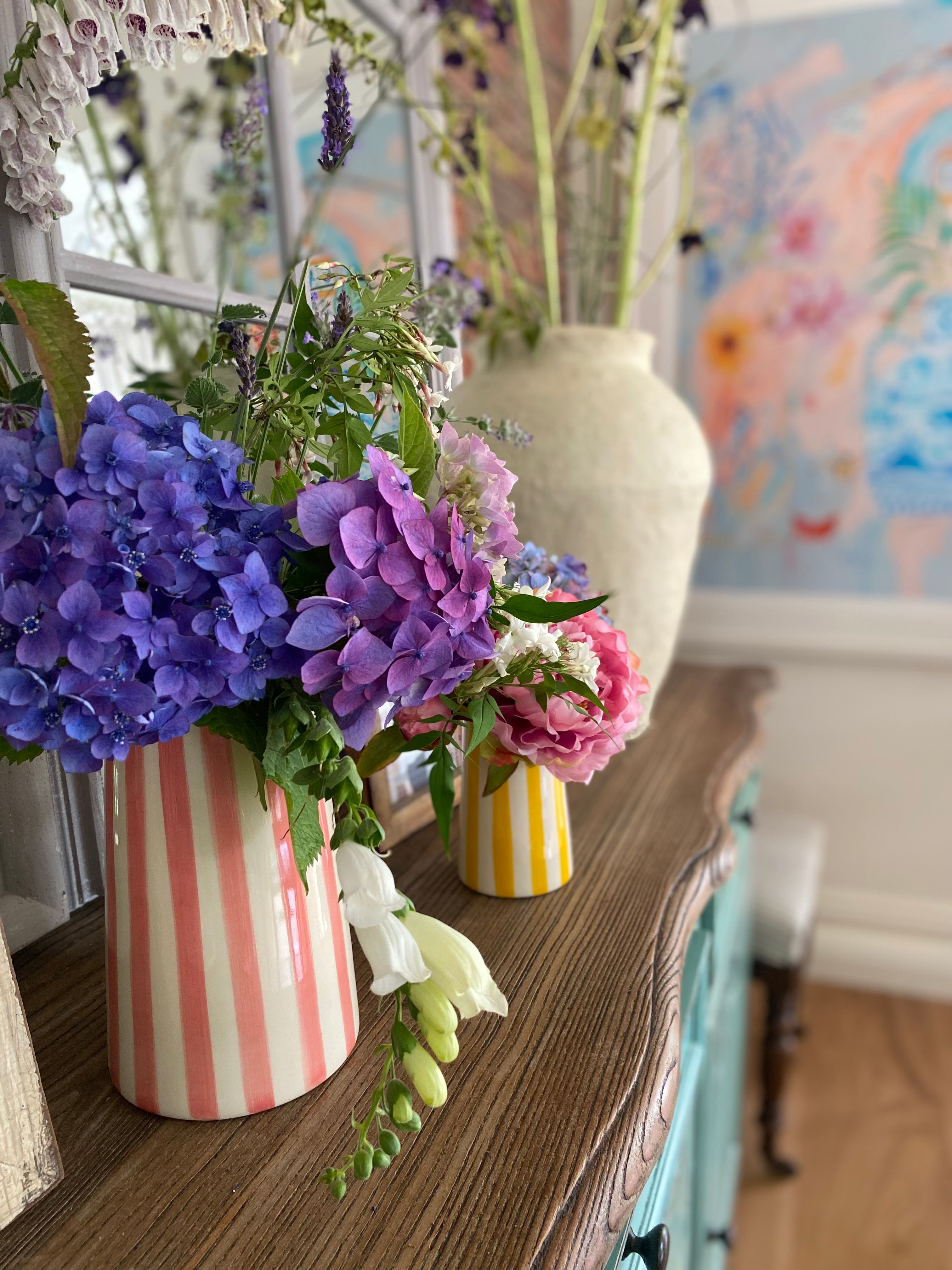 Pink and yellow striped pitcher and jugs filled with colourful flowers on a sideboard in a colourful room
