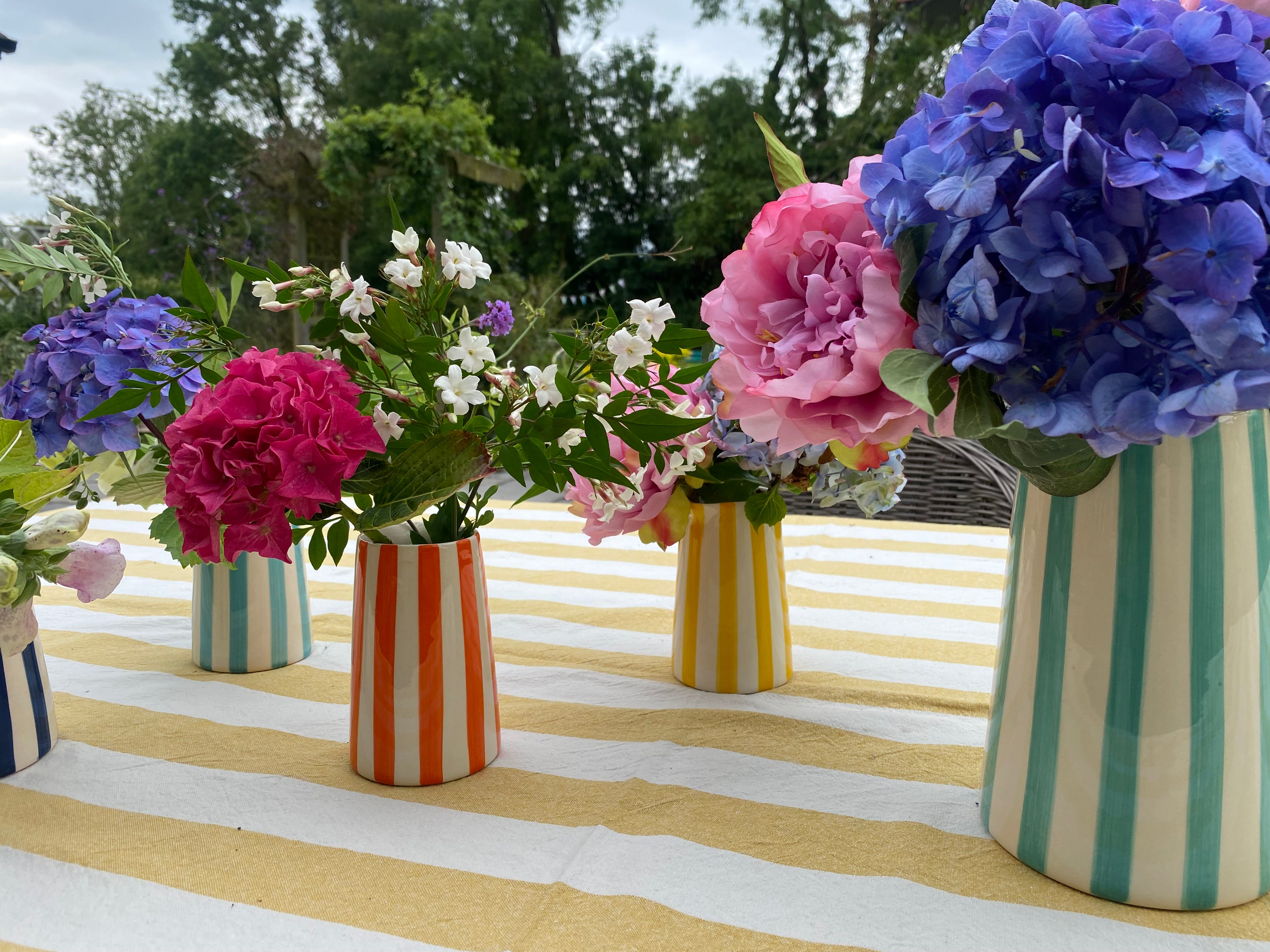 Striped pitchers filled with colourful flowers on a table with yellow and white linen tablecloth in sunny outdoor setting