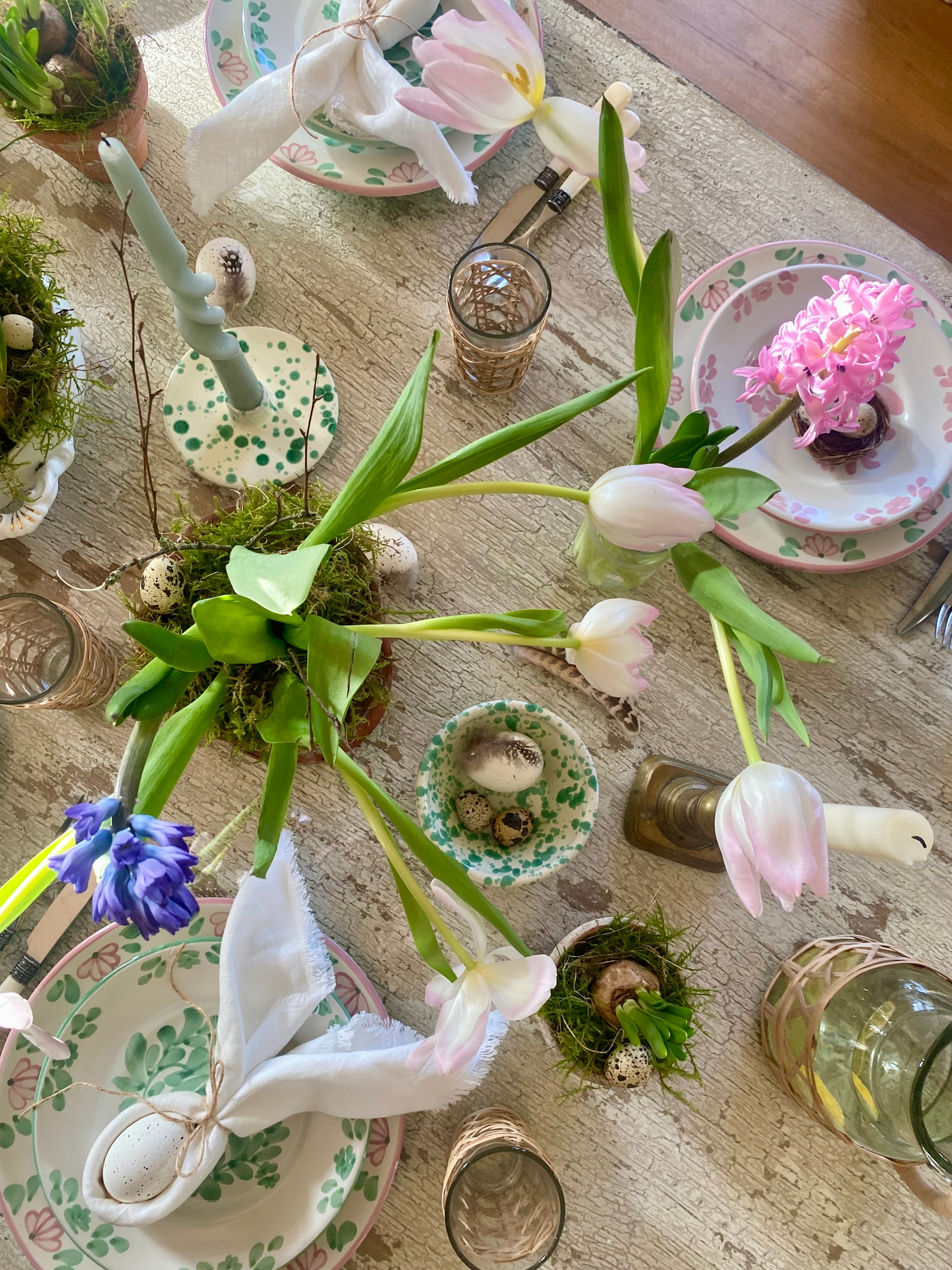 Overhead shot of Spring Easter table with green splatter bowls, candle holders, mossy daffodil bulbs, hyacinths, tulips ,napkins tied into bunny ears