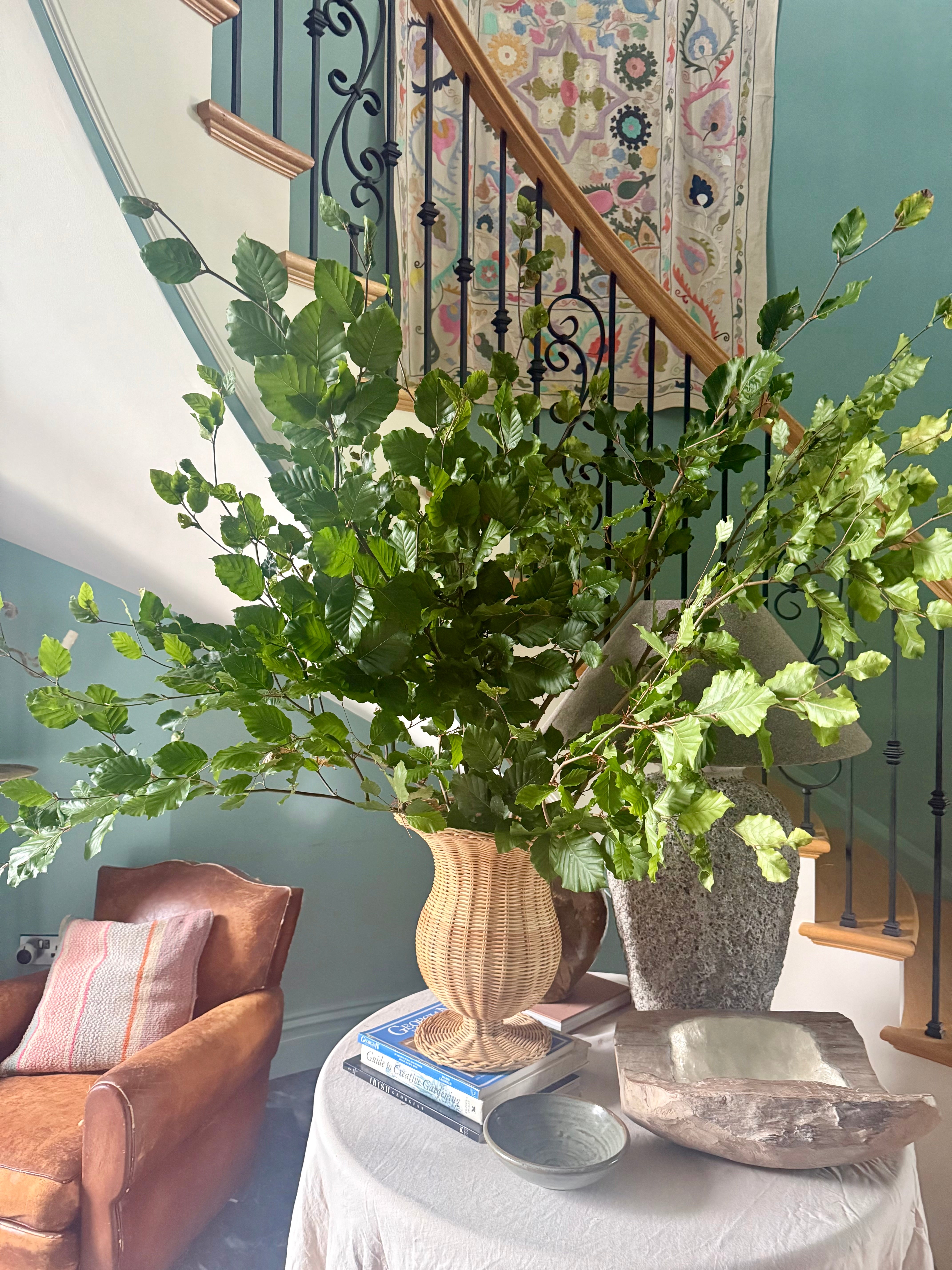Decorative indoor setting with a large green branches in a rattan urn on a table, surrounded by books and a sculptural bowl.
