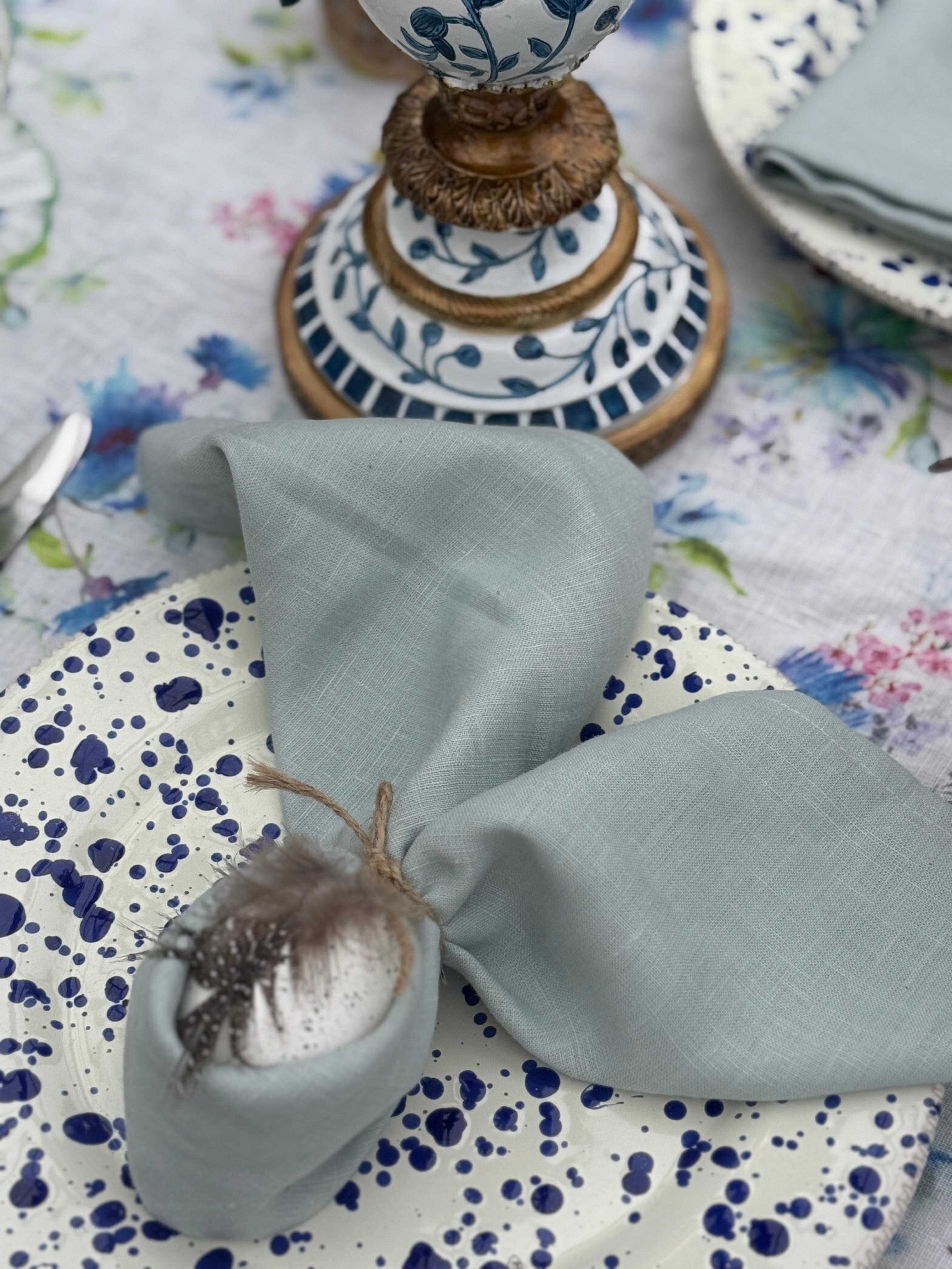 A set of dusty blue linen napkins on a table with a floral tablecloth, displayed alongside other tableware.