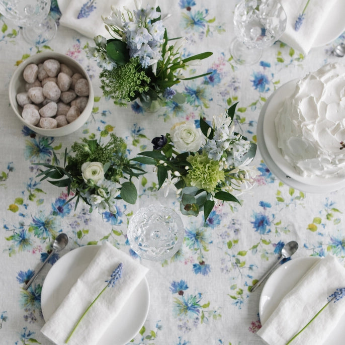 Overhead shot of floral tablecloth with white linen napkins, white cake, sugared almonds, and green flowers