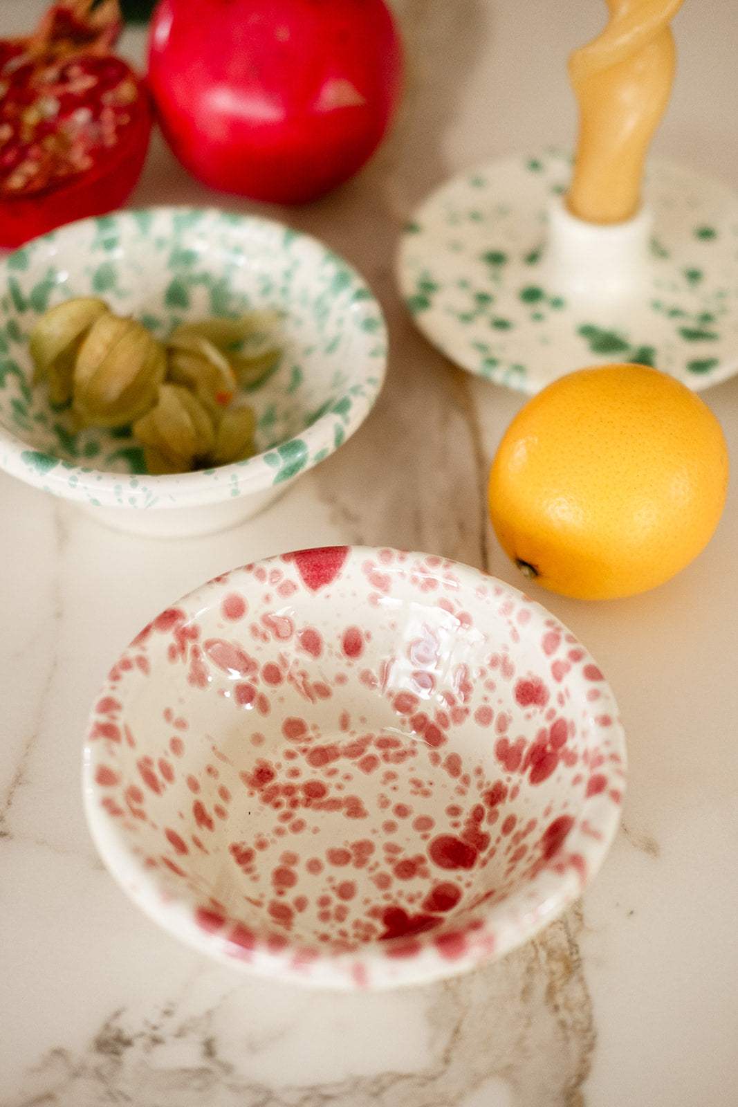 Close up of red splatter bowl, green splatter bowl and green splatter candleholder with apricots and fruits on white marble surface