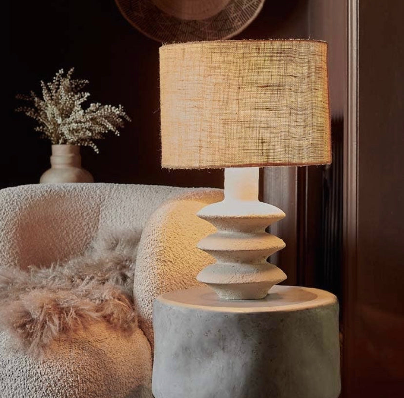 The Akito lamp with a creamy hued textured base and a jute lampshade, placed next to a white chair with a fluffy blanket on it, in a moody brown cozy home setting.