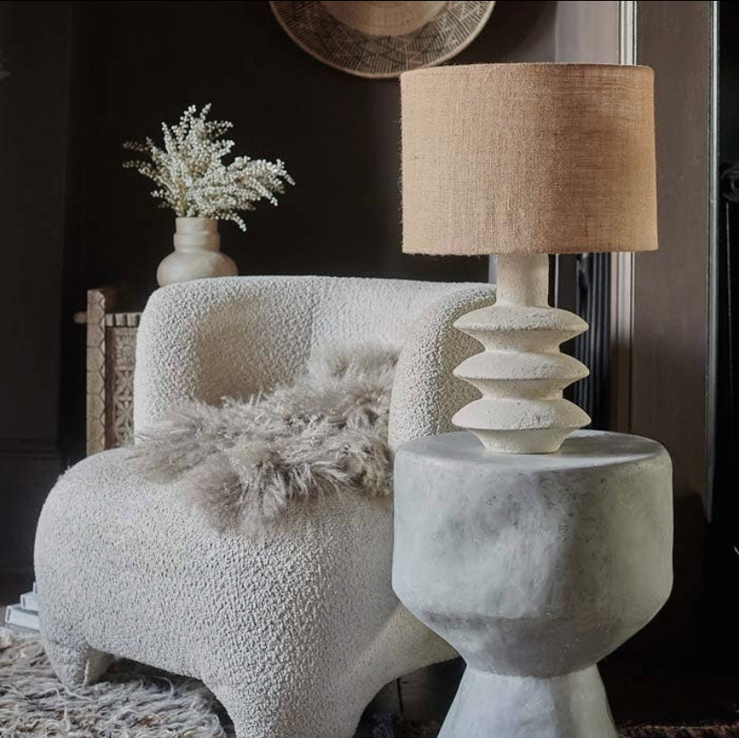 The Akito lamp with a creamy hued textured base and a jute lampshade, placed next to a white chair with a fluffy blanket on it, in a textural home setting in daylight
