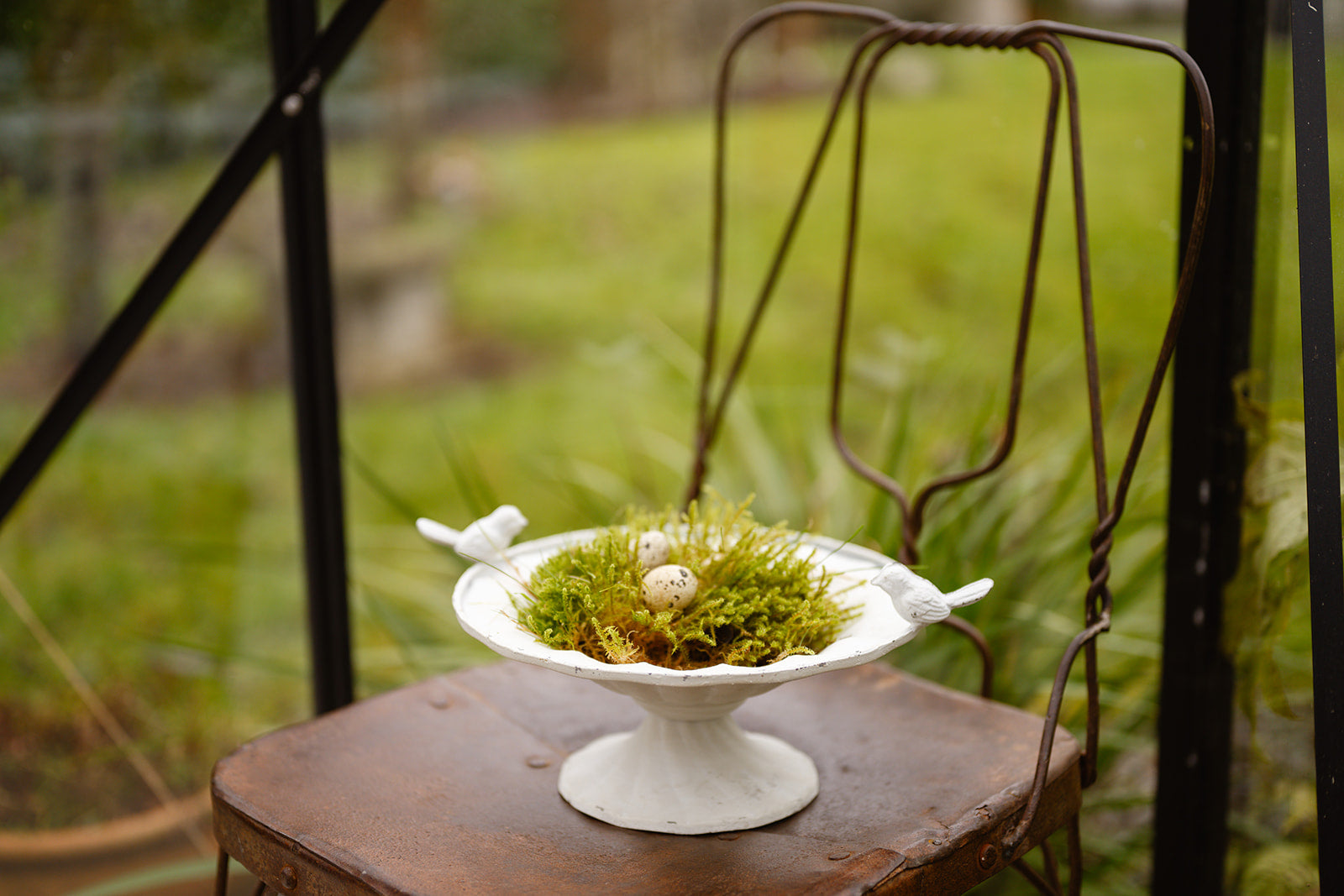 Decorative arrangement with moss and eggs on a white pedestal in front of a rustic chair outdoors.