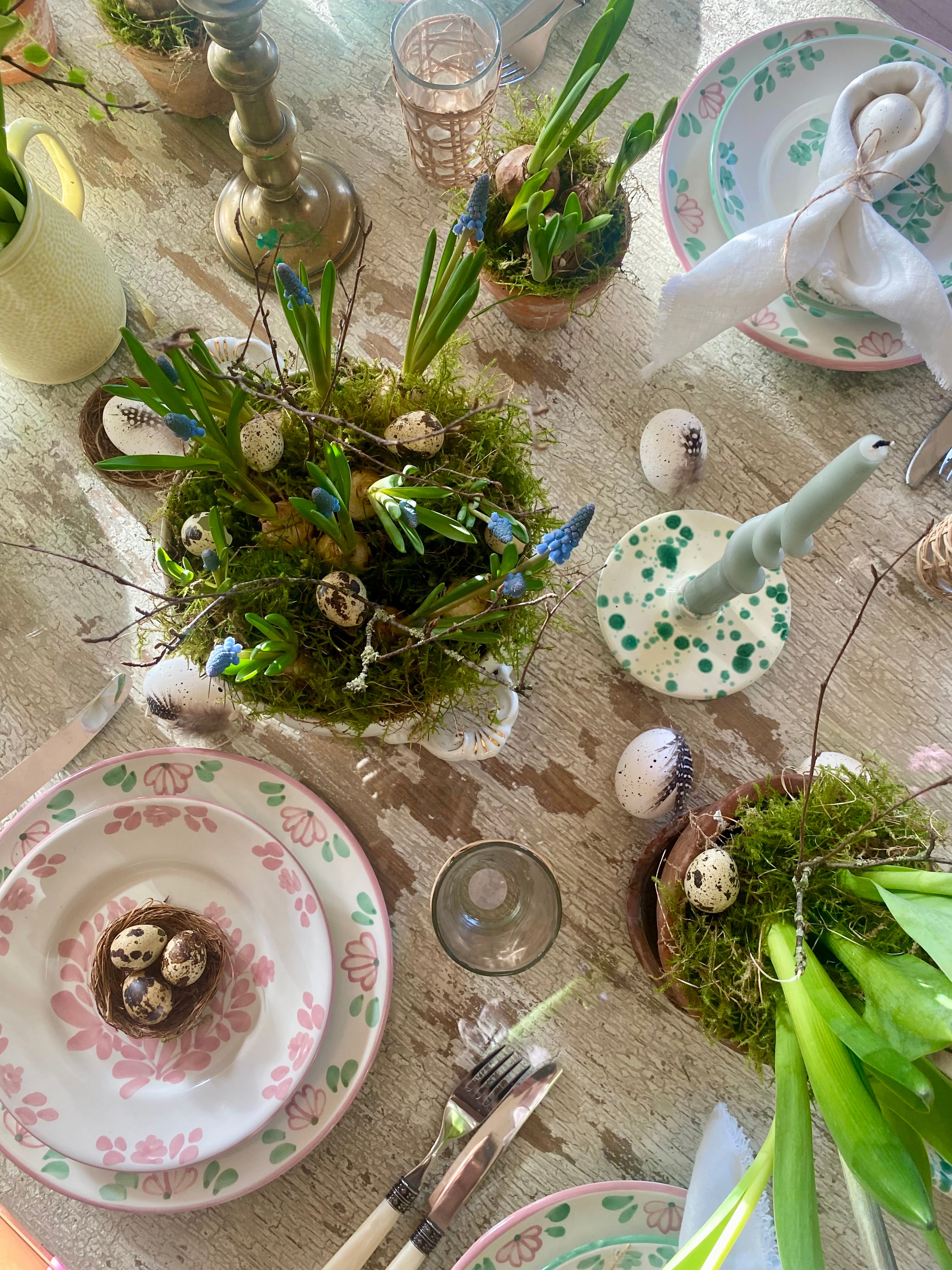 Overhead shot of Spring Easter table with green splatter bowls, candle holders, mossy daffodil bulbs, hyacinths, tulips ,napkins tied into bunny ears, quail eggs, muscari