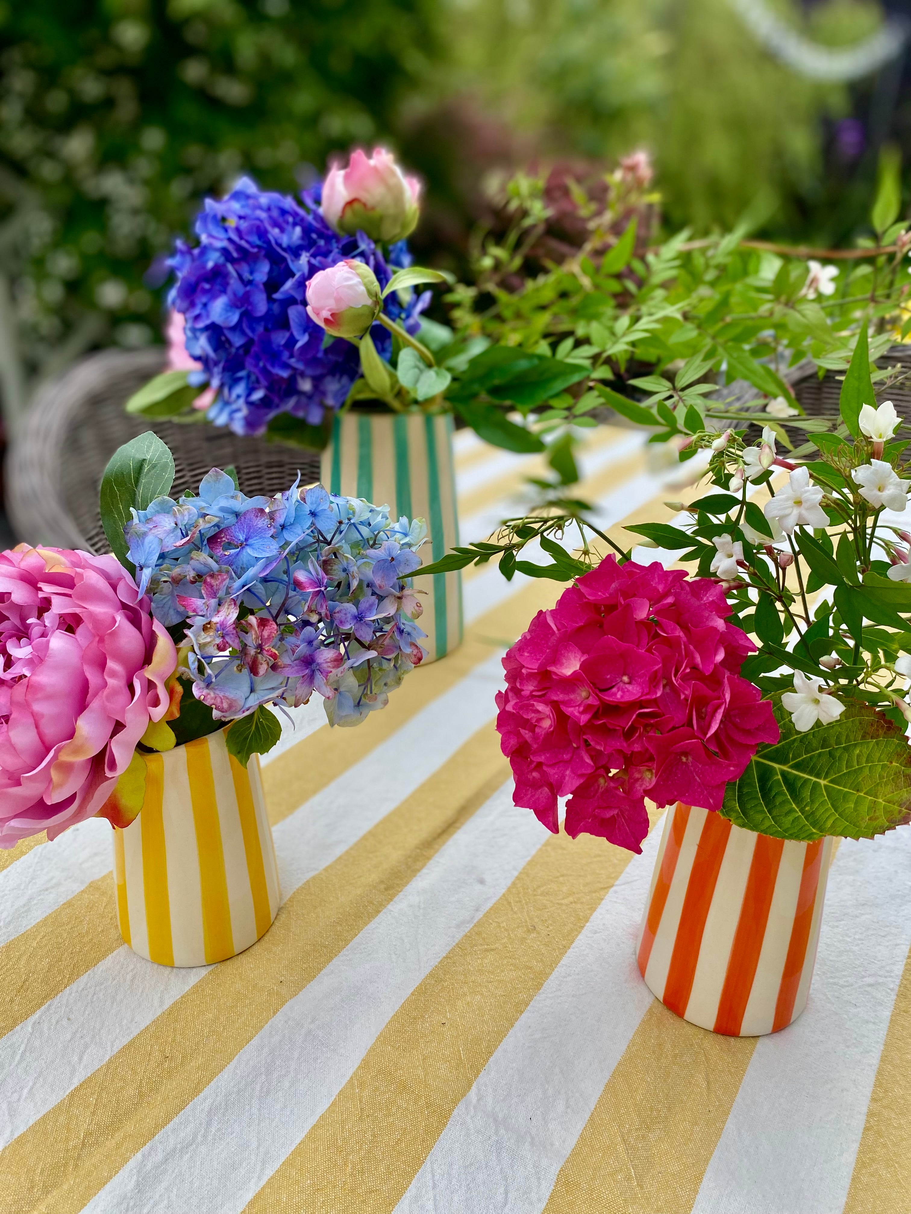 Striped pitchers filled with colourful flowers on a table with yellow and white linen tablecloth in sunny outdoor setting