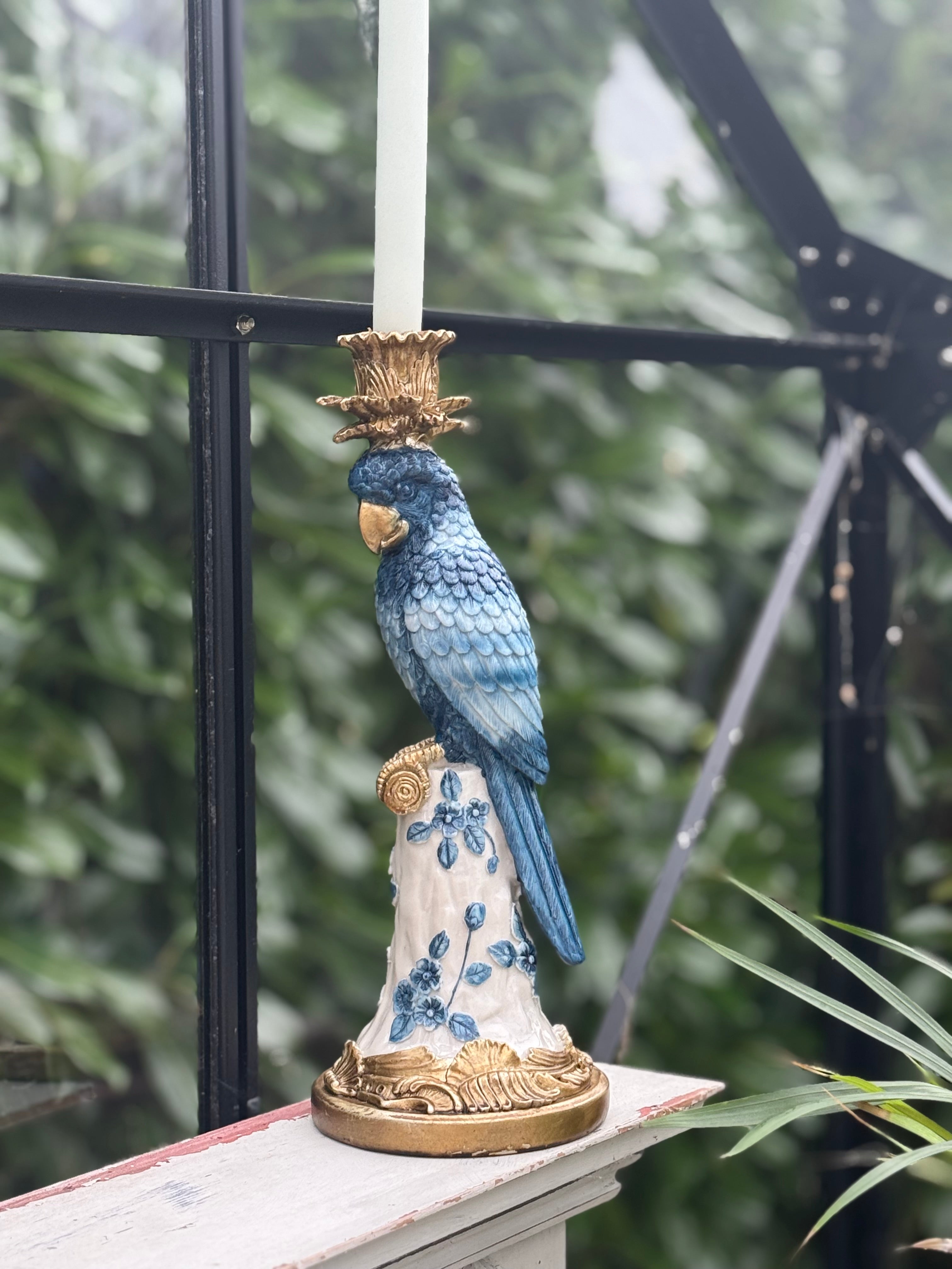 This image features a whimsical and colorful blue parrot candle holder. The candle holder is designed with a detailed and vibrant depiction of a parrot in mid-flight, landing on a ornate, gold-accented perch.