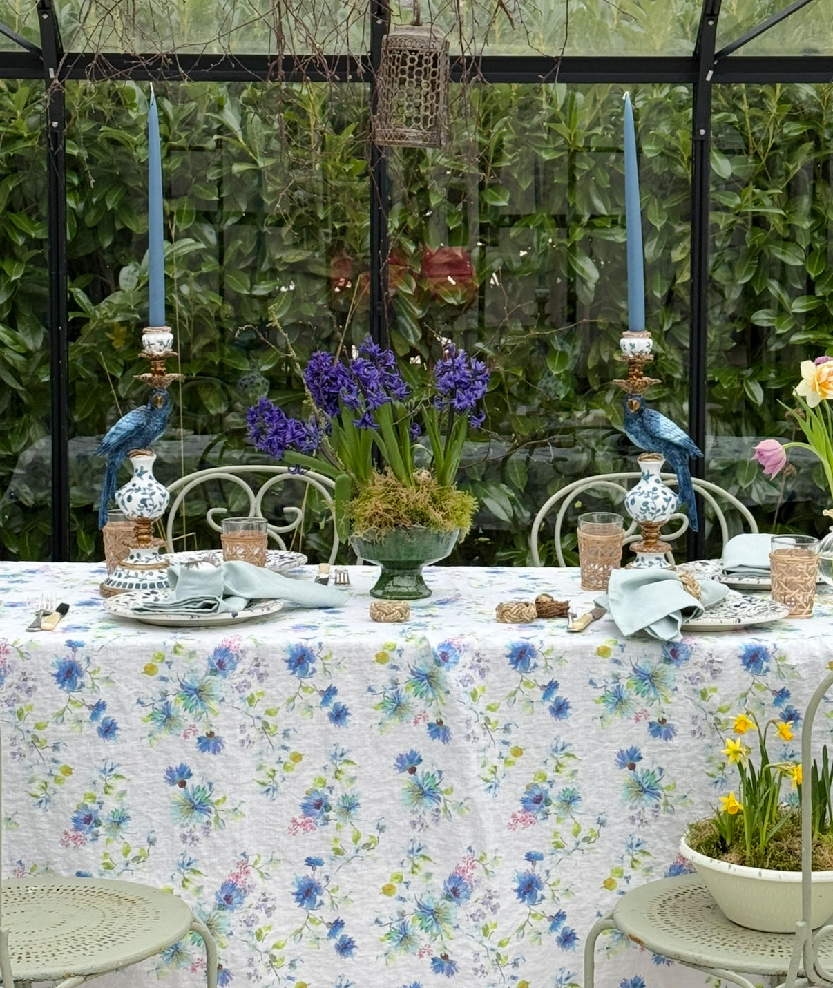 Table setting with white linen floral tablecloth with blue and green tones with chinoiserie blue parrot candle holders, blue candles in a glass house
