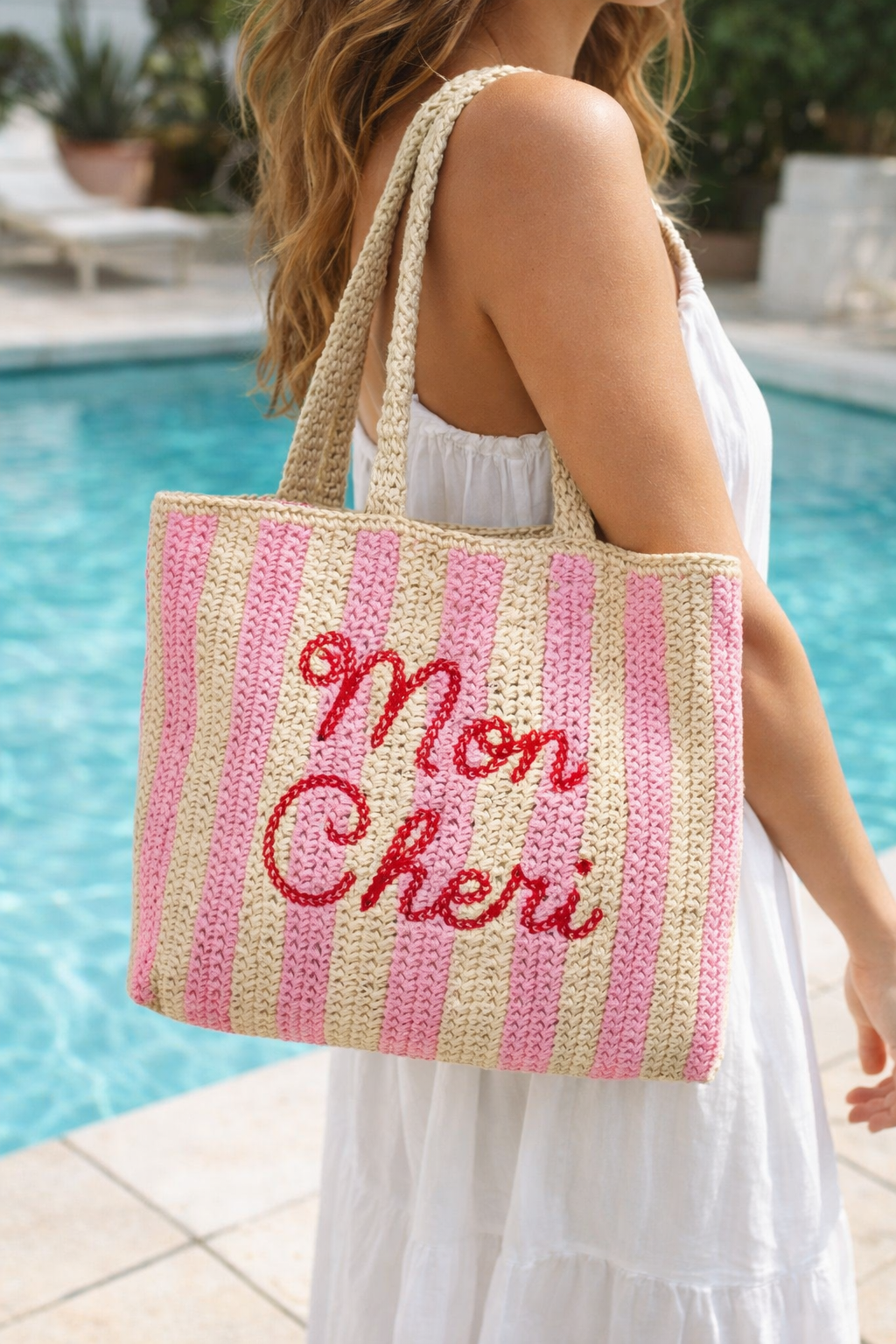 Woman in white sun dress carrying a raffia pink striped beach bag with 'Mon Cheri' embroidered text by a pool.