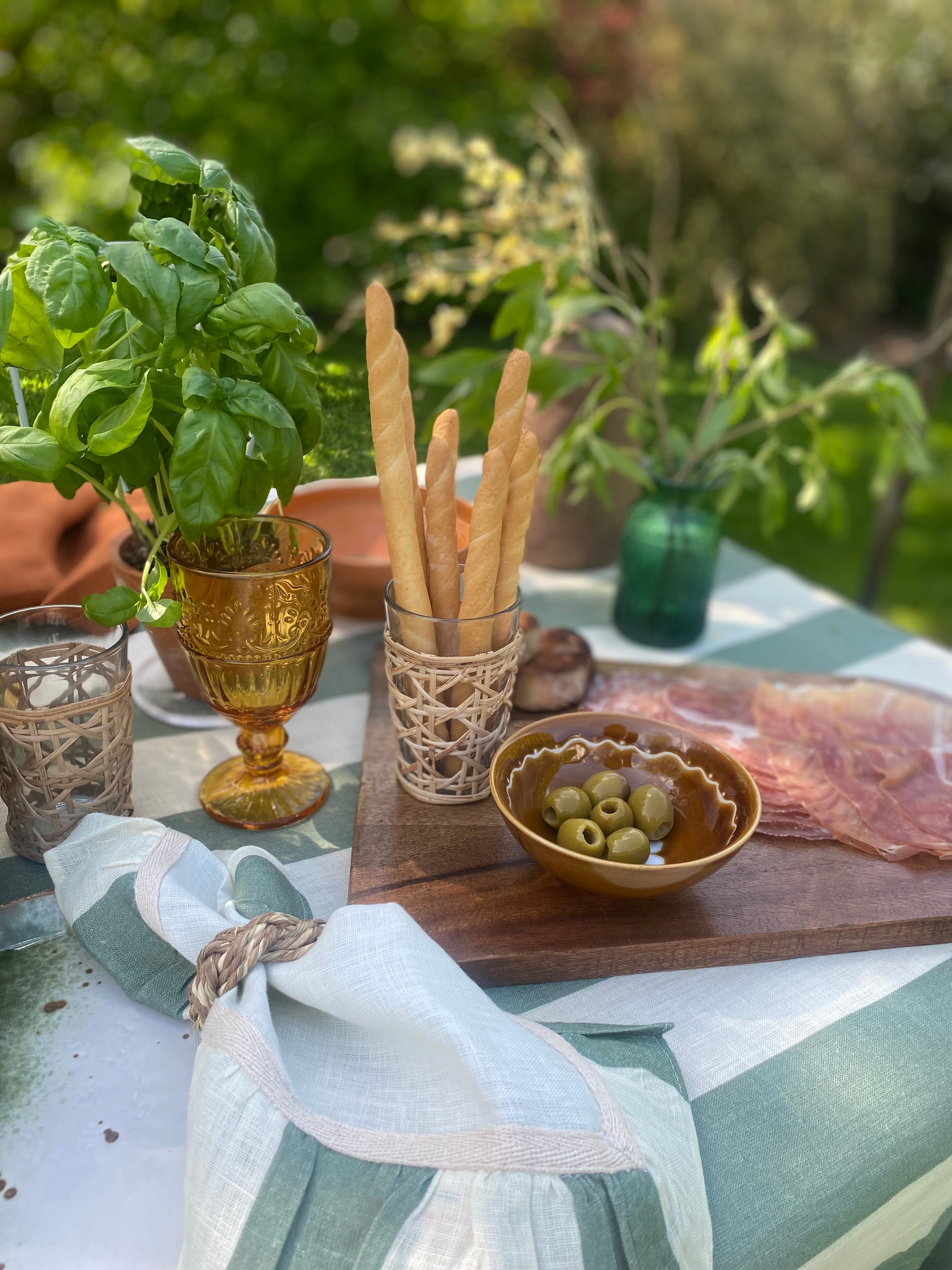Outdoor sunny table setting with mango wood board with charcuterie and bowl of olives, breadsticks, green and white linen tablecloth