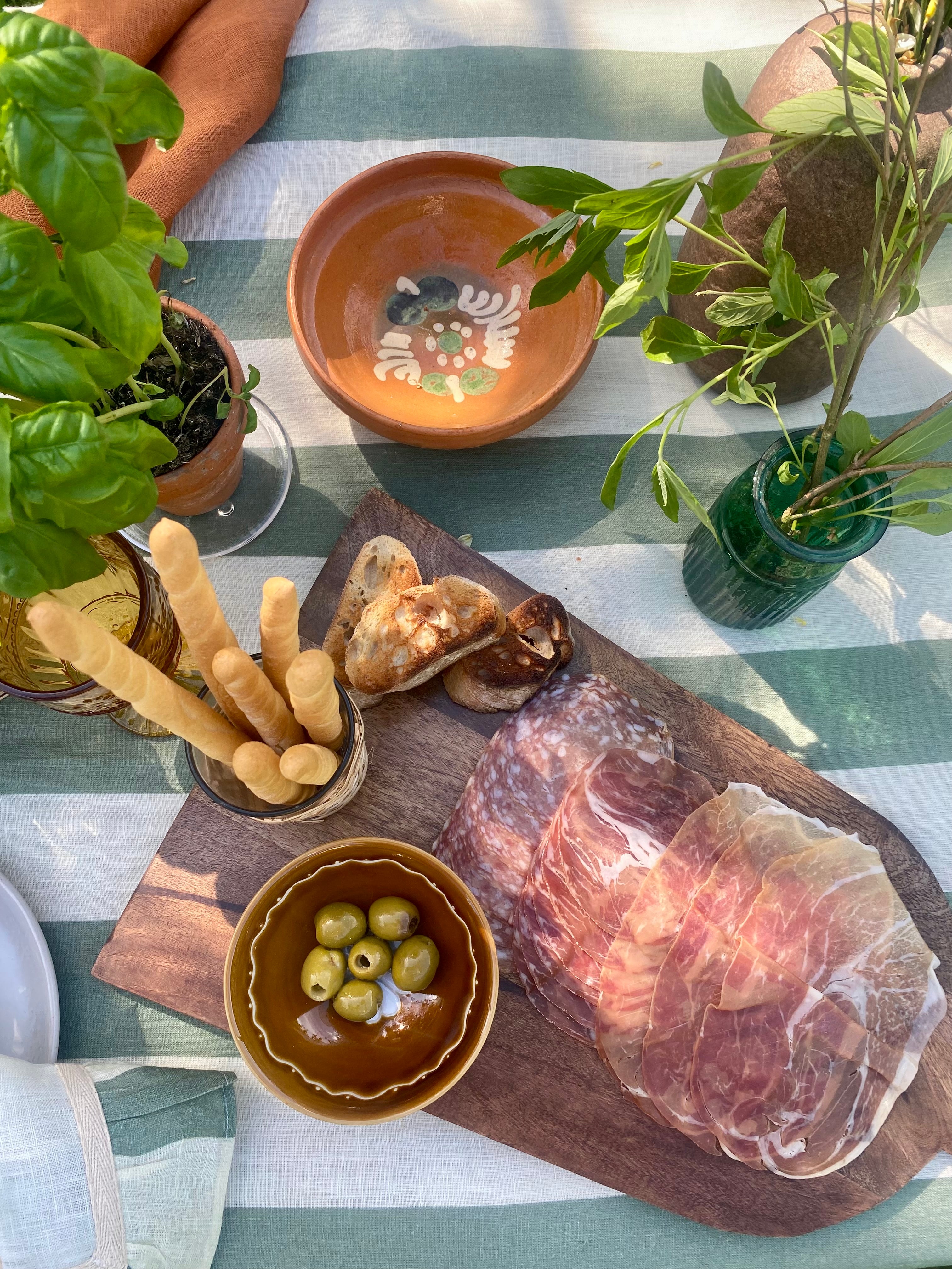Table with cured meats on a wooden board, bread sticks, and olives on green and white linen tablecloth.