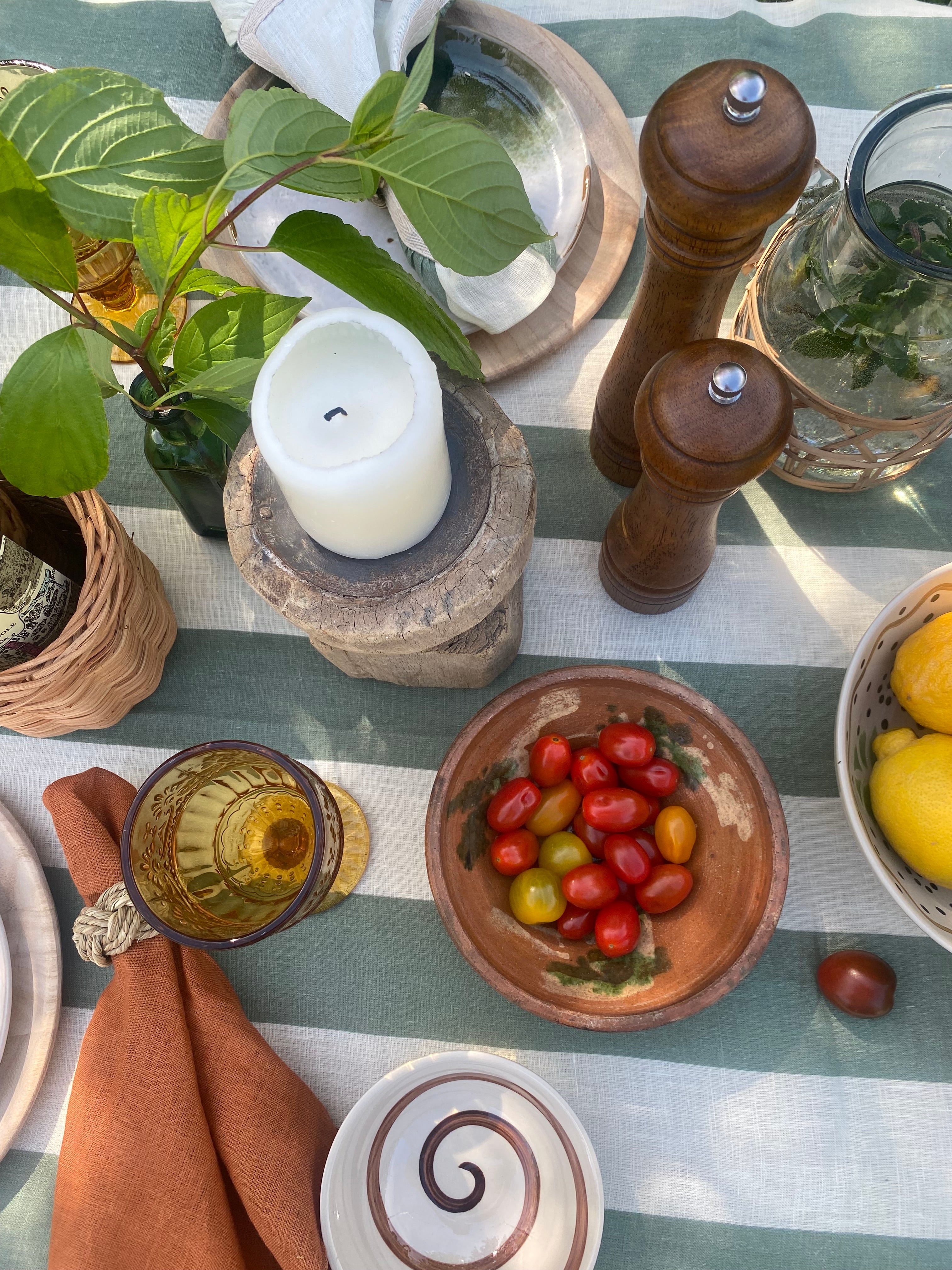 Overhead view of a table with green and white striped linen tablecloth, bowl of tomatoes, terracotta napkin, ochre glassware , candle and other elements