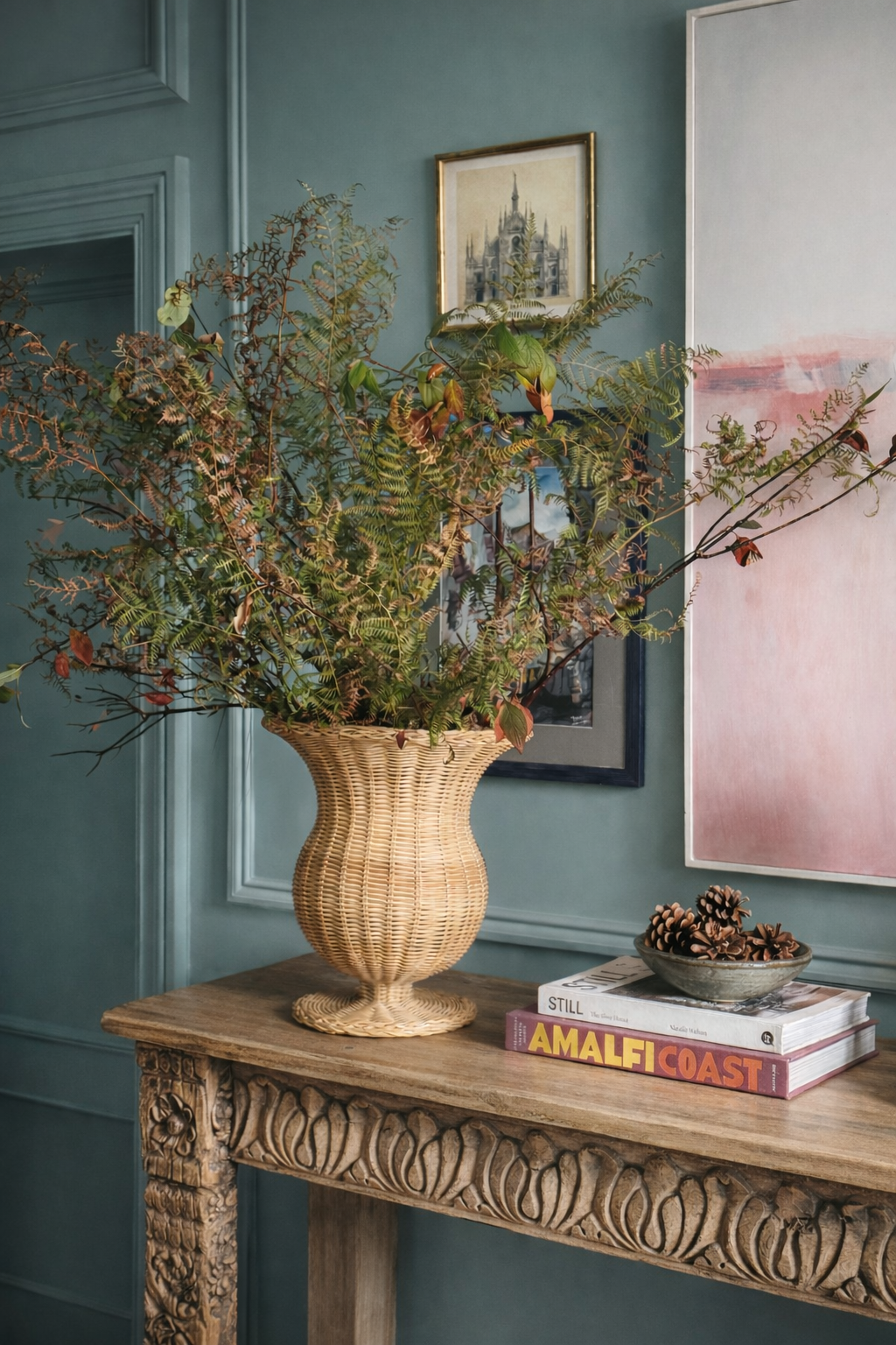 Rattan urn with rustic branches on carved indian console table against a bright teal panelled wall with a mix of artwork.