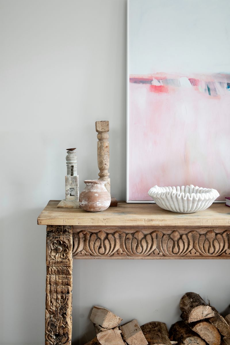 Carved Indian wooden console with fire logs underneath styled with reclaimed wood candleholders next to vintage marble ruffled bowl with pink abstract art on the wall above.