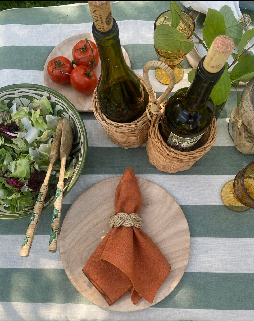 An overhead styled table setting with a green and white striped tablecloth, a bowl of mixed salad with wooden spoons, two wine bottles in a woven rattan holder, a plate of red tomatoes, and a round wooden plate with terracotta-coloured linen napkin tied with a seagrass napkin ring.