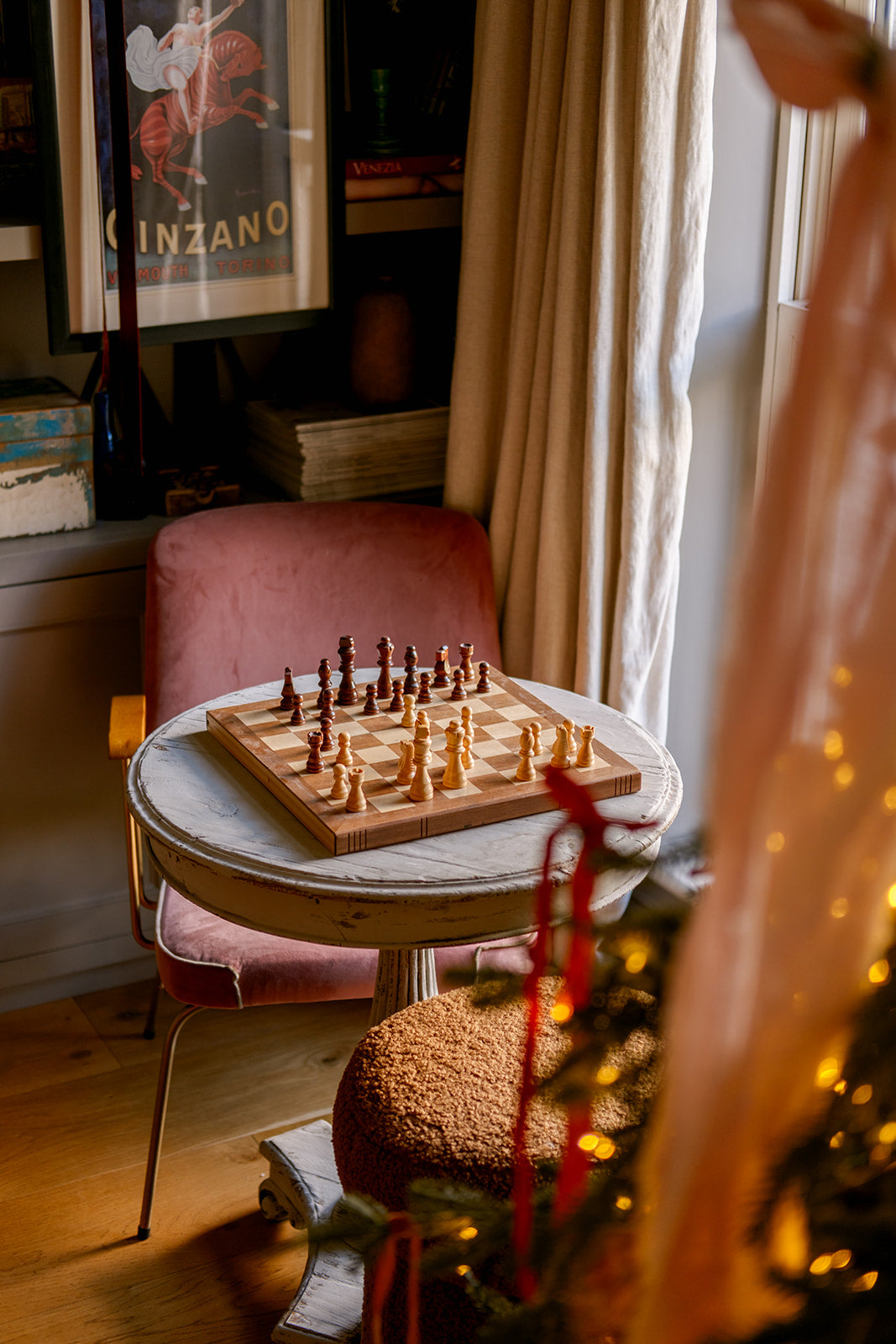 A wooden chessboard with chess pieces set on a small round table in a cozy room with a pink chair, curtains, and warm holiday lights.