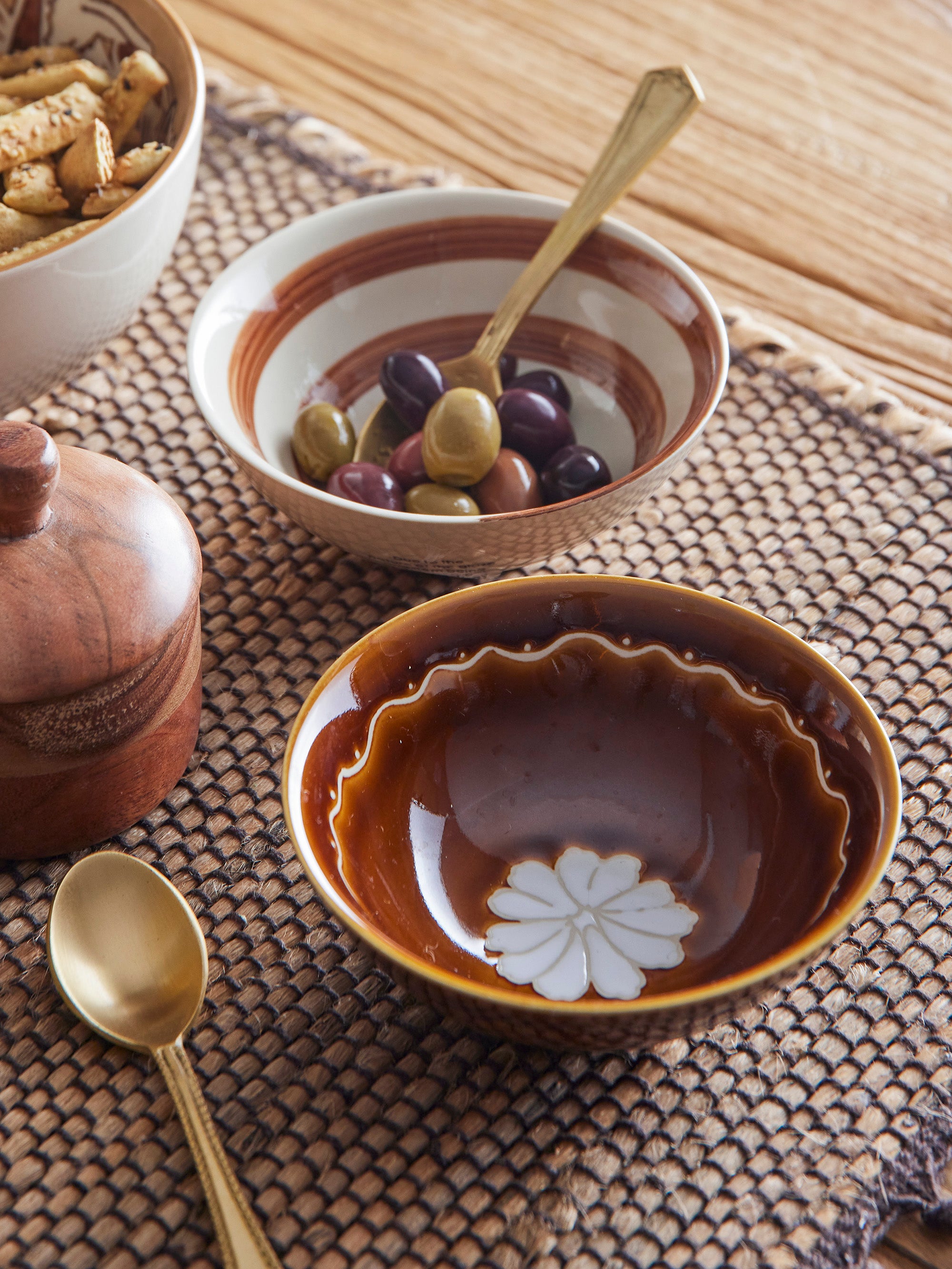 Close up of handpainted cream and brown bowls won woven place mat next to other bowls and cutleryth olives