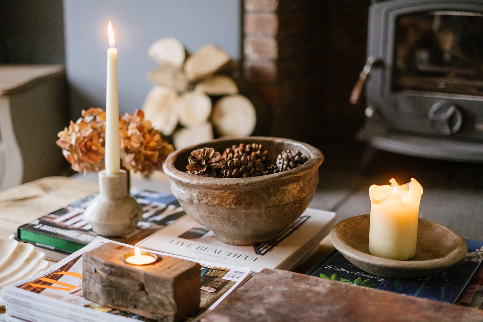 Candlelit scene with a grey papier-mache bowl of pinecones, candles, and magazines on a coffee table.
