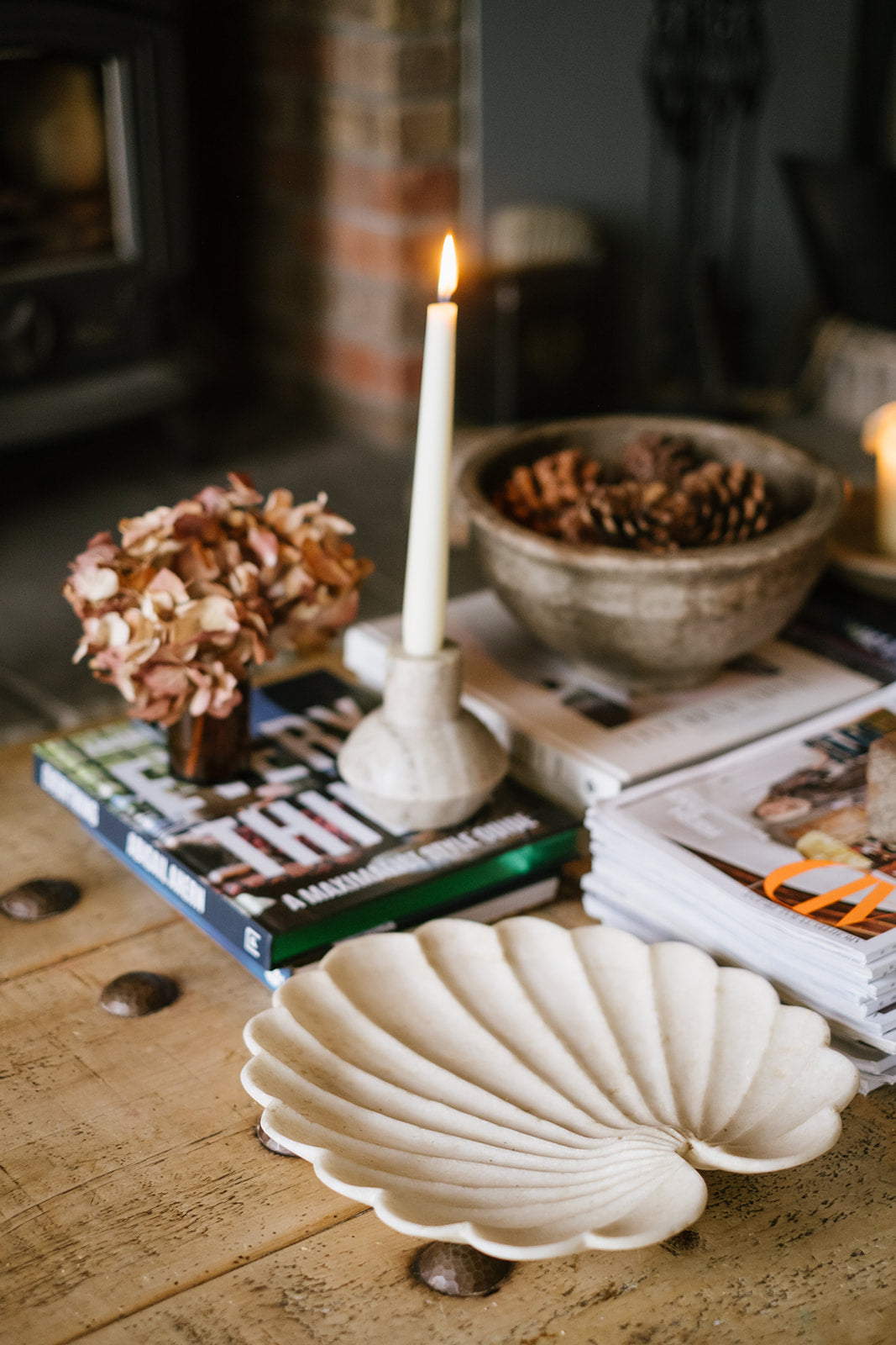 View of rustic wood coffee table setting with marble shell plate, bowl of pinecones, candles lighting, stacks of books