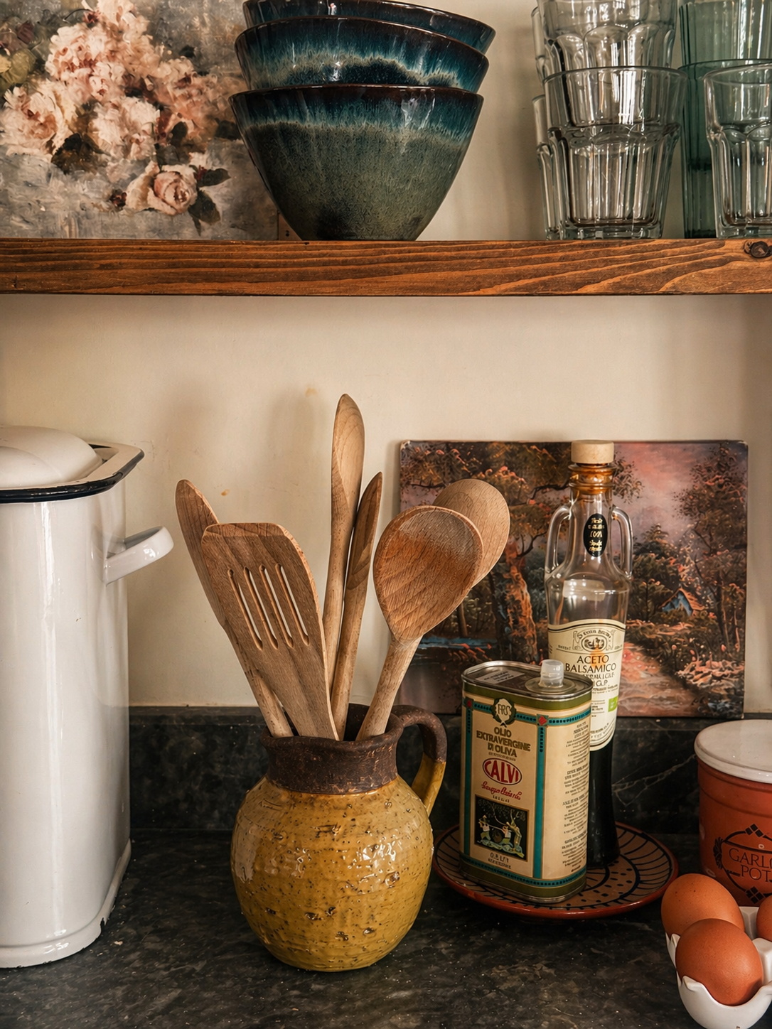 Wooden utensils in a yellow glazed terracotta jug on a kitchen counter next to bottles of olive oils, balsamic vinegar, eggs in a ceramic holder, and vintage white metal bread bin items in the background.