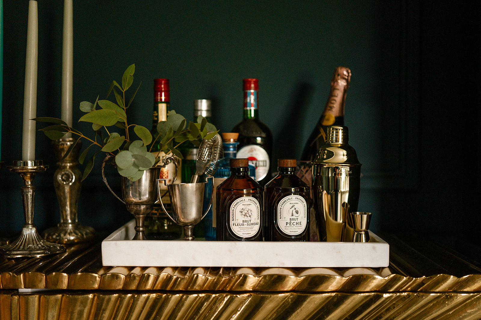 Bottles of spirits and champagne, gold cocktail maker, old silver trophy cups with greenery marble tray with a dark background