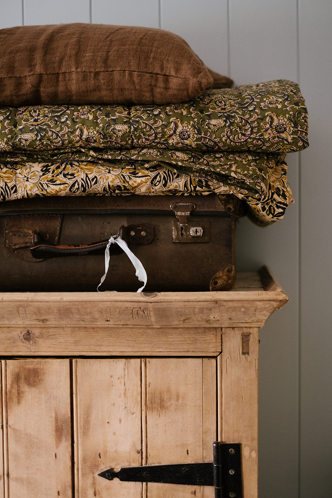 A throw with floral print and tassels draped over a wooden piece of furniture, with cushions and a vintage suitcase in the background.