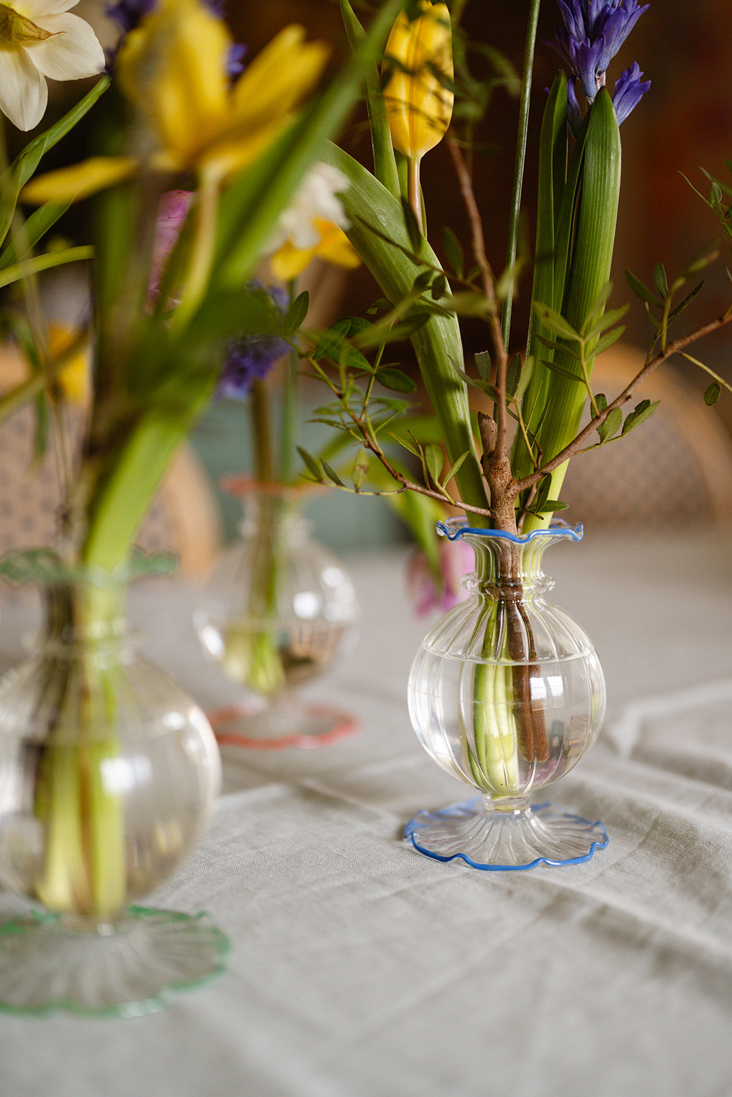 Clear glass vases with green stems and flowers on a tablecloth.