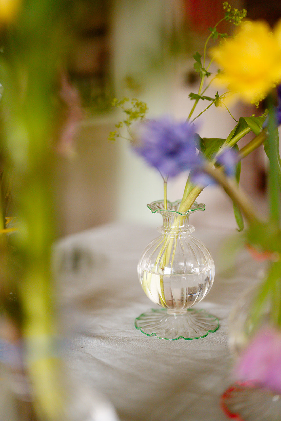 Small clear glass vase with water and green plant stem on a blurred floral background