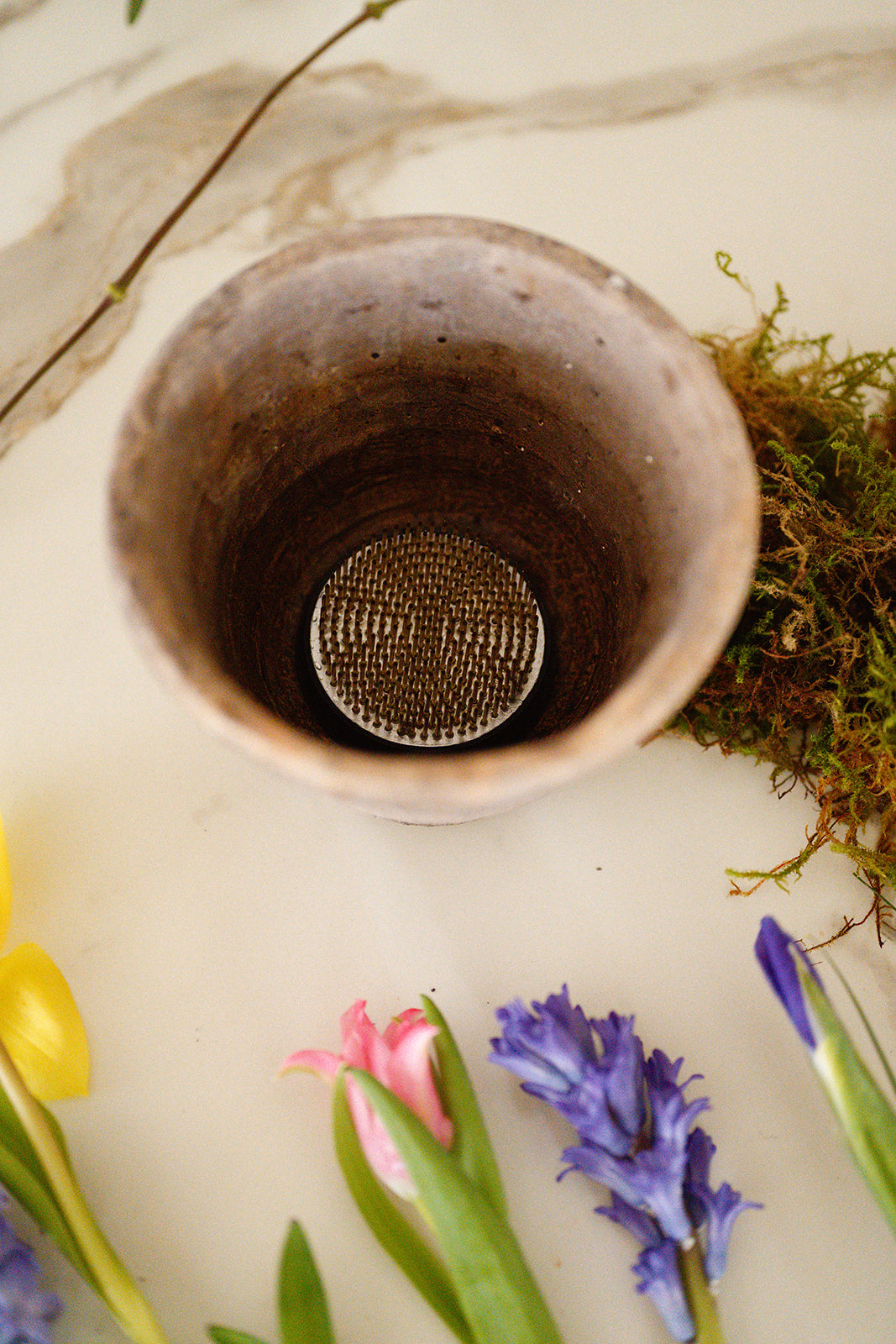 Detail shot of flower frog in bottom of aged pot with spring flowers laid out on white surface