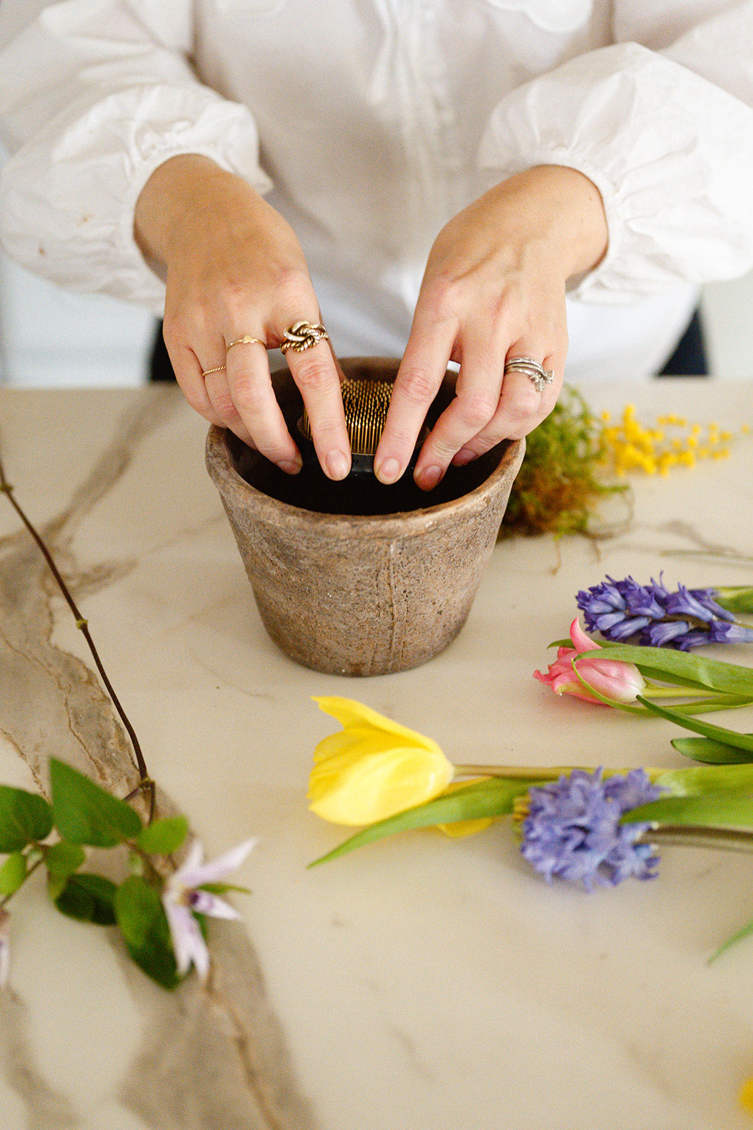 Amber + Willow placing flower frog into aged pot on white surface with spring flowers laid out