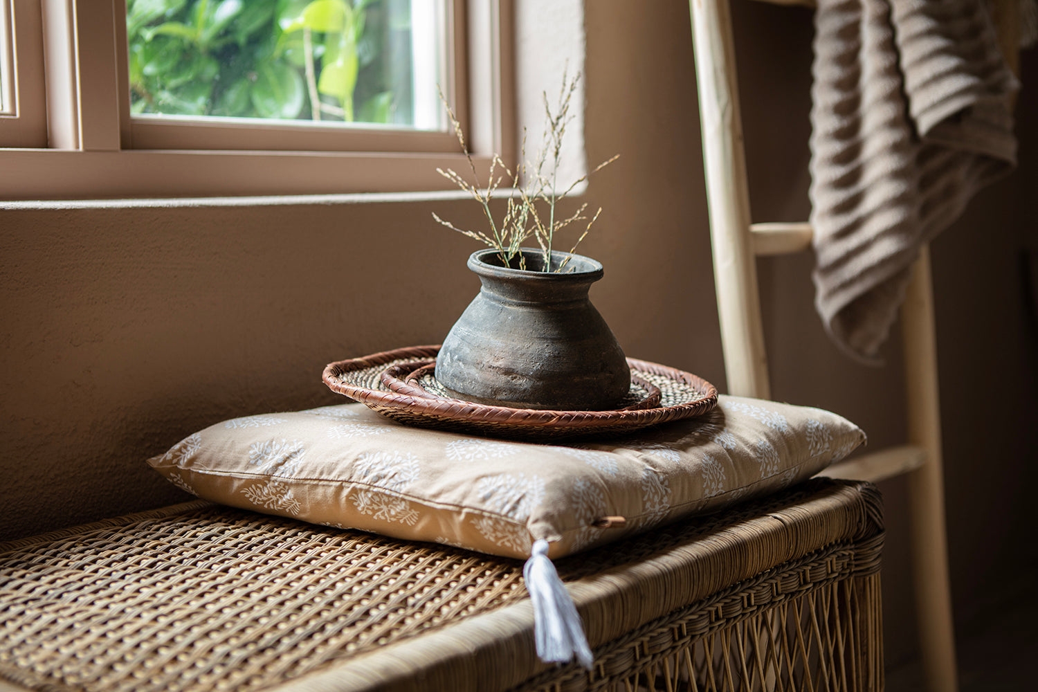 Decorative pot with dried plants on a pillow on a wicker bench by a window.