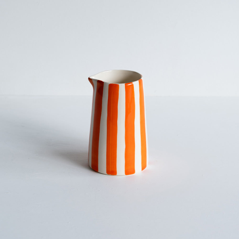 A handmade ceramic creamer jug with orange and white stripes on a white background.