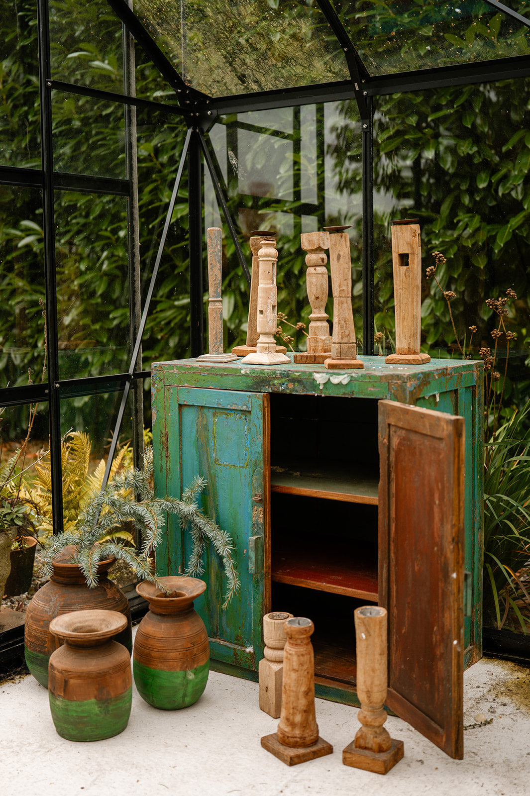 Vintage green cabinet with open doors, surrounded by wooden vases and pots in a greenhouse setting.
