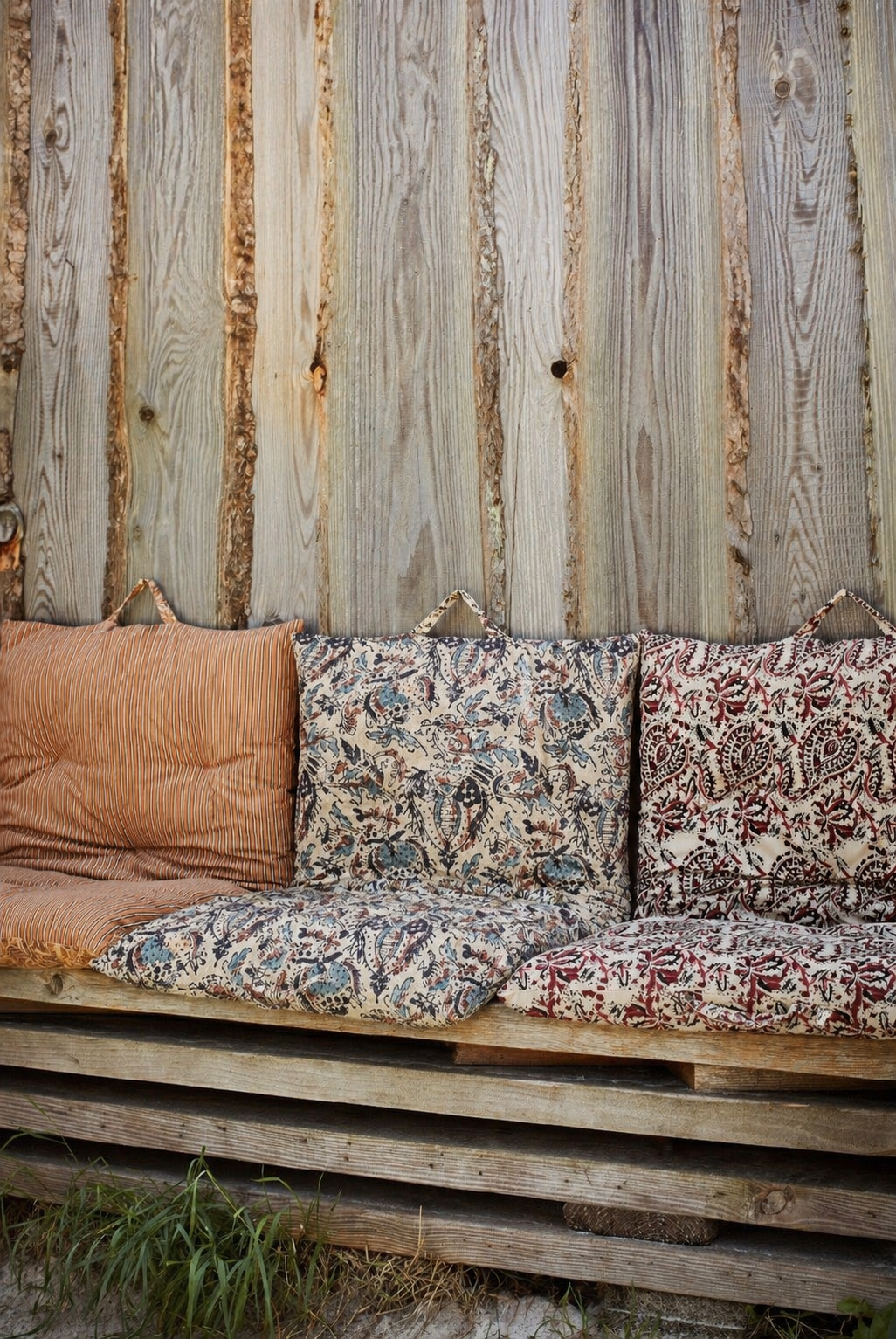 Pallet bench with mismatched block print patterned cushions against a rough hewn wood fence