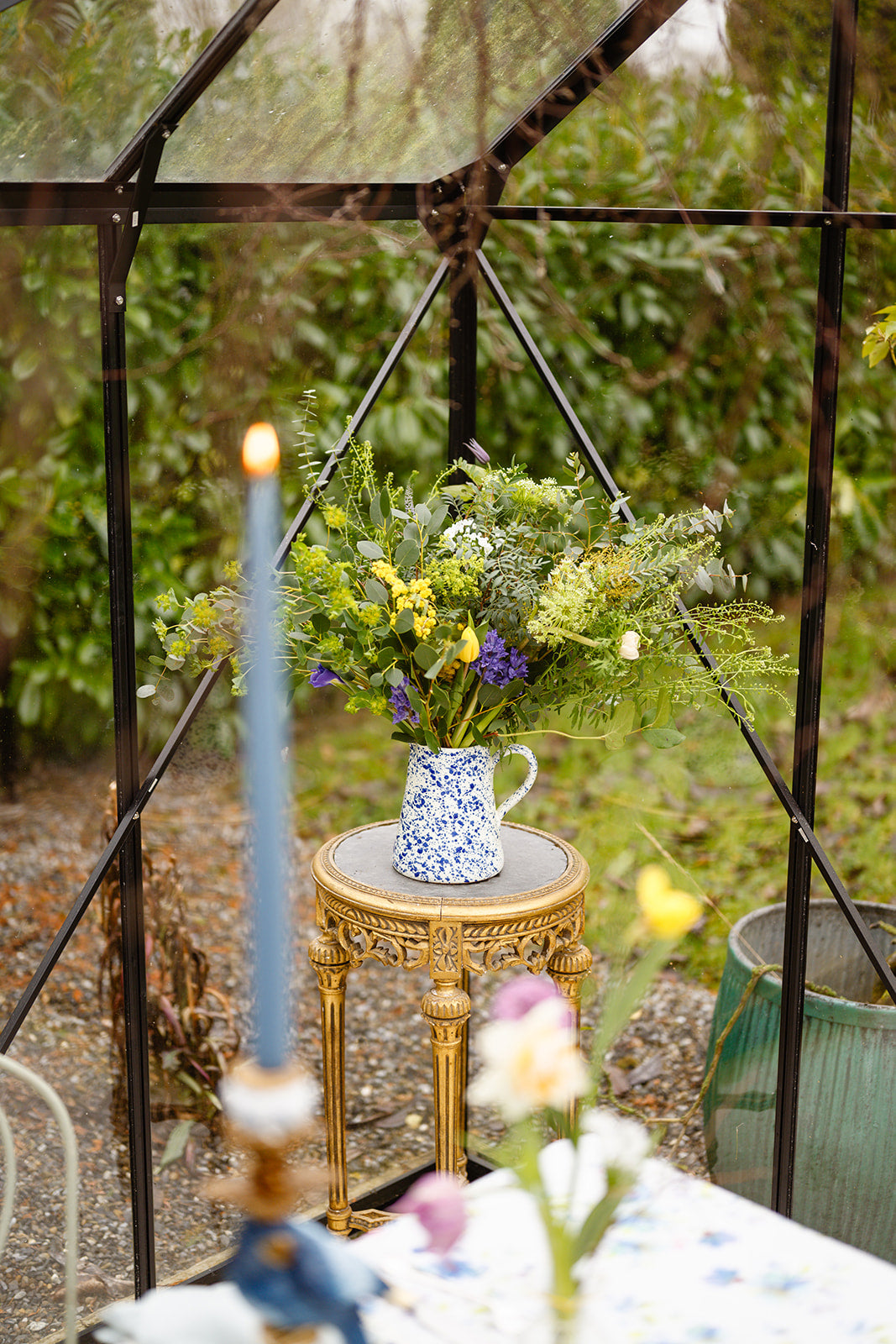 Glass house setting with black frame with a gold round table with blue splatter jug filled with flowers in garden setting