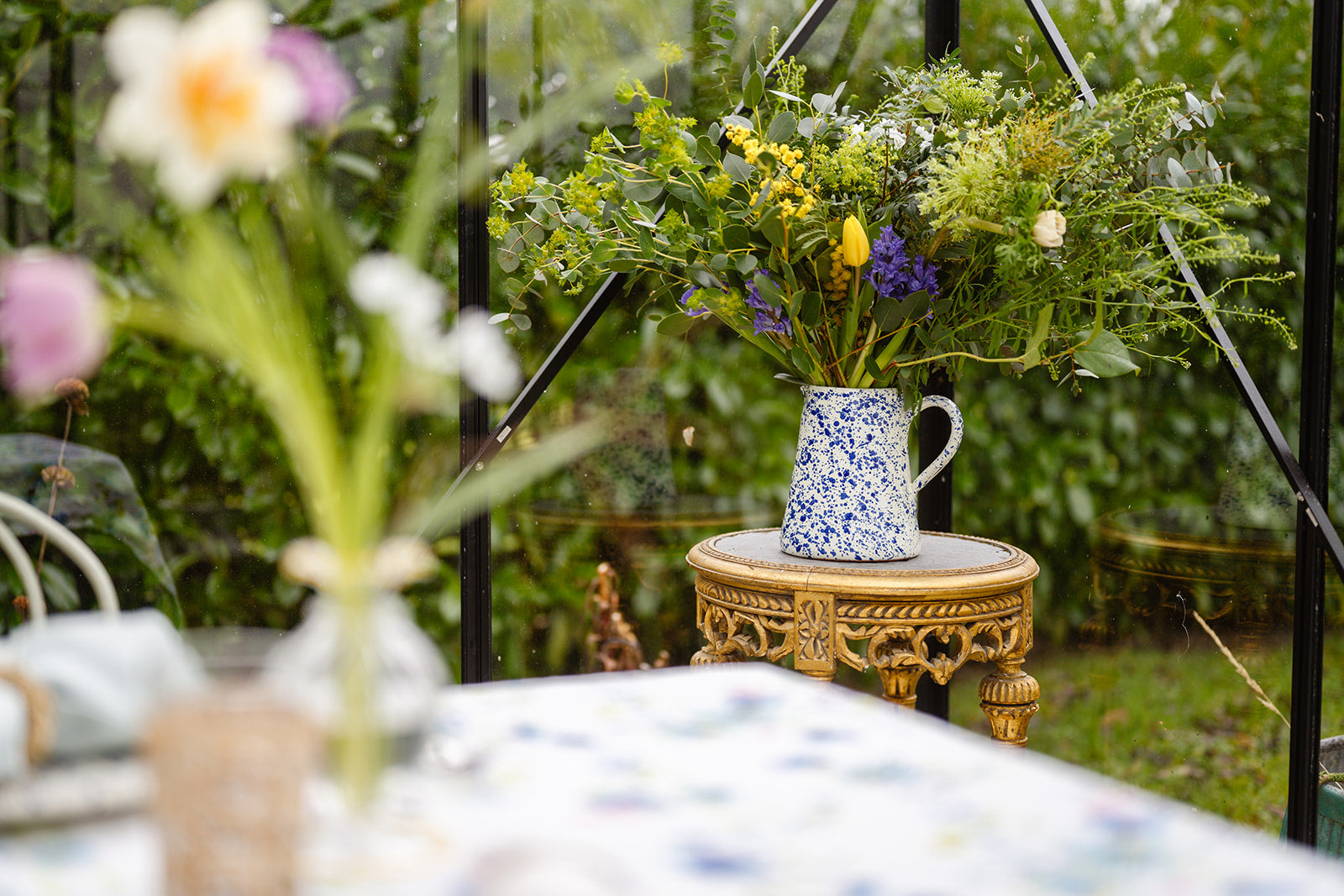 Glass house setting with black frame with a small round antique gold table with blue splatter jug filled with flowers in garden setting