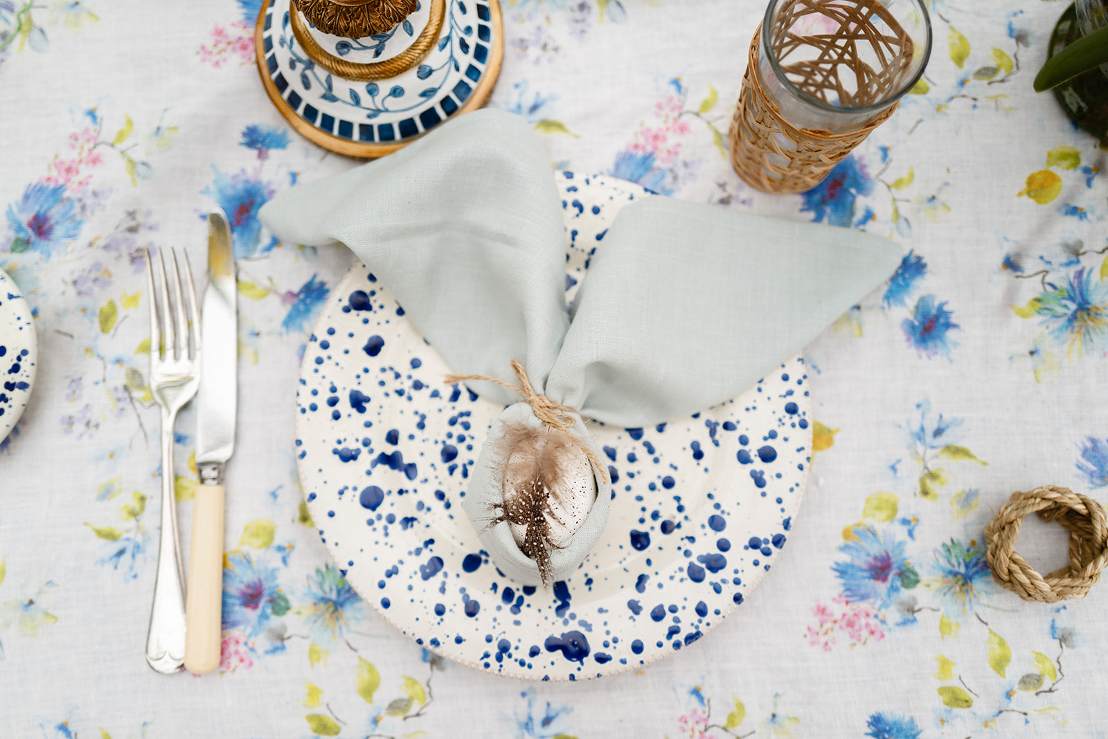 Table setting with blue splatter plates, duck egg napkin tied into bunny ears with a decorative egg , and floral tablecloth.