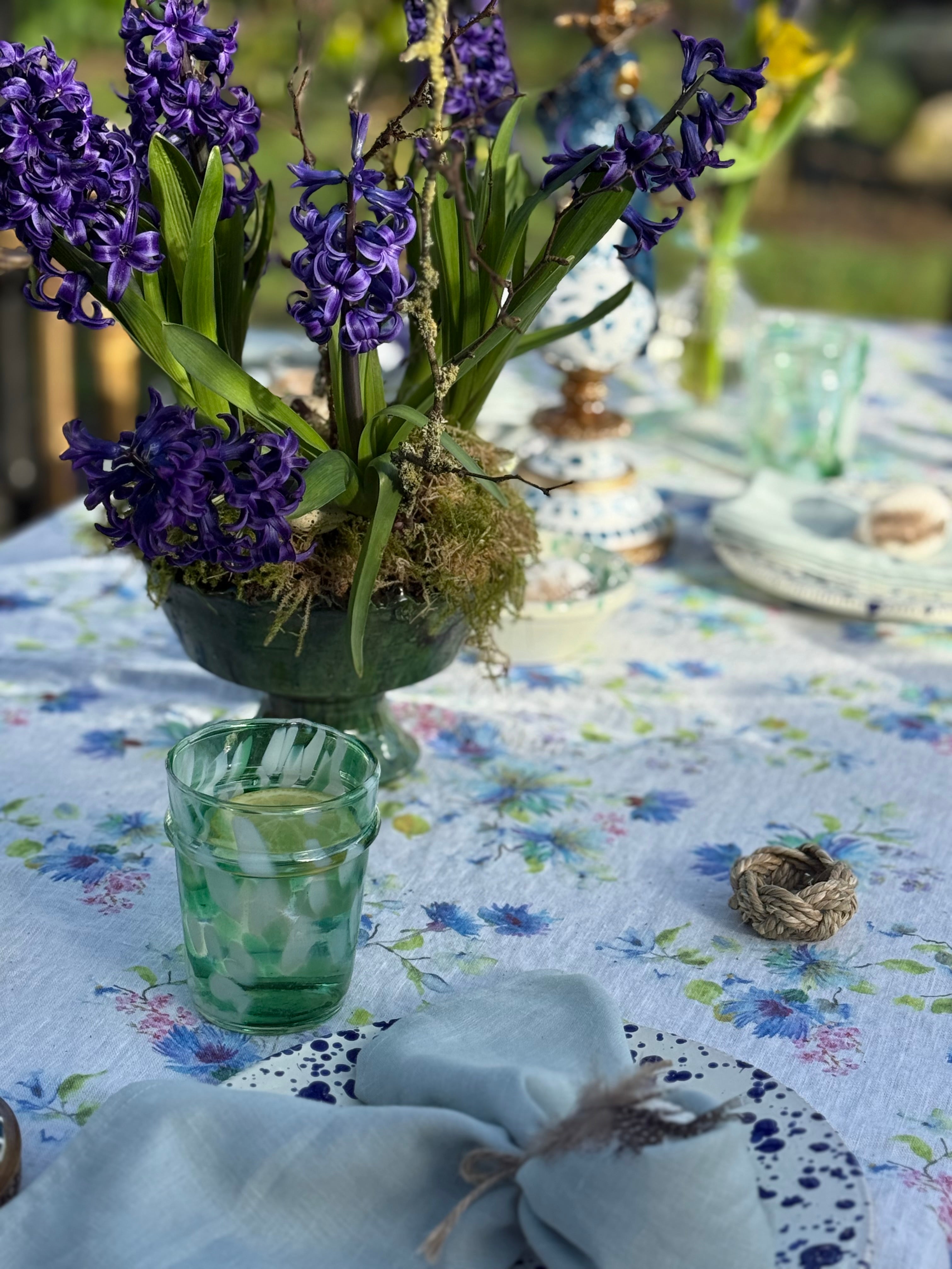 Spring garden table with purple hyacinths in green bowl with moss on floral linen tablecloth, duck egg blue bunny ear napkins, sea grass napkins and green and white splatter glasses