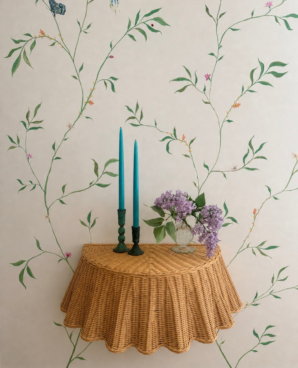 Floating rattan skirted console table with verdigris candle holders with turquoise candles and vase of fresh lilac against a hand painted botanical wall mural