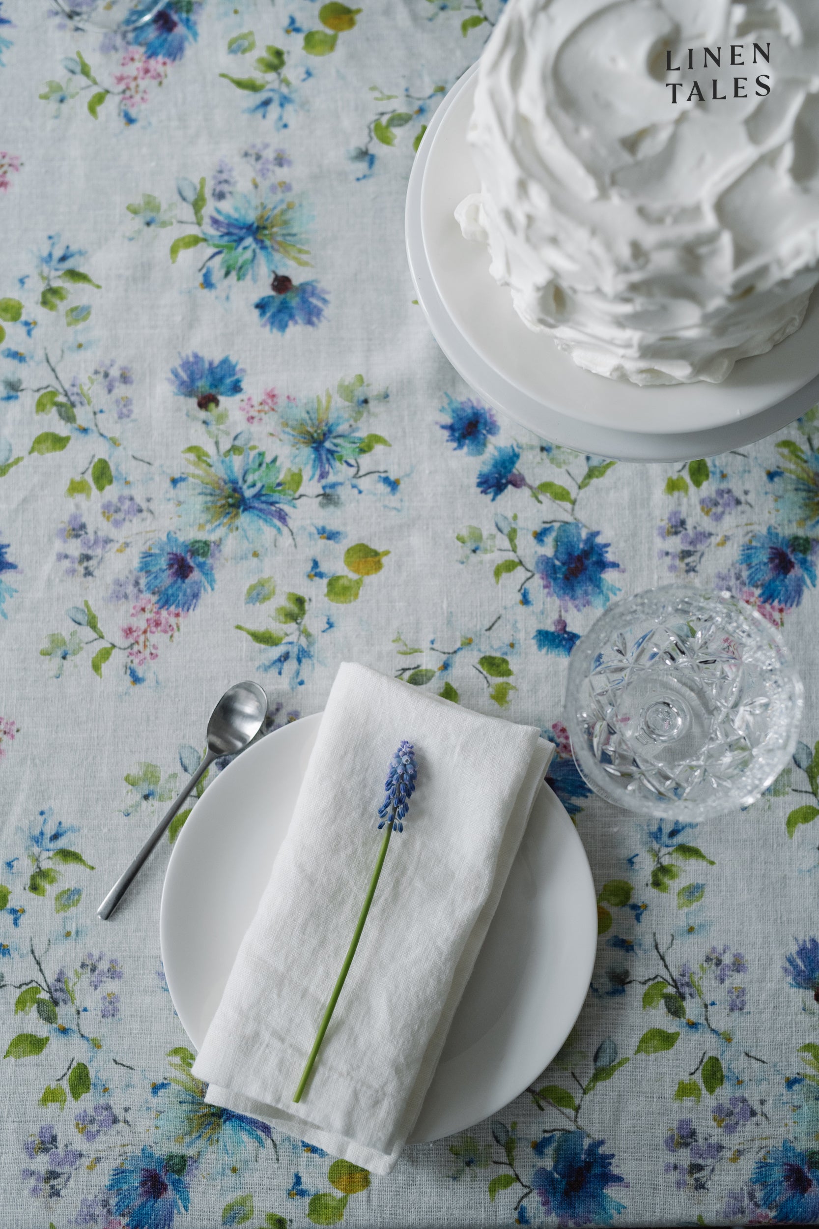 A white linen tablecloth with a delicate floral pattern in blue and green, displayed on a table set with a white plate, silver cutlery, and a glass. A cake with white frosting is also visible in the background.