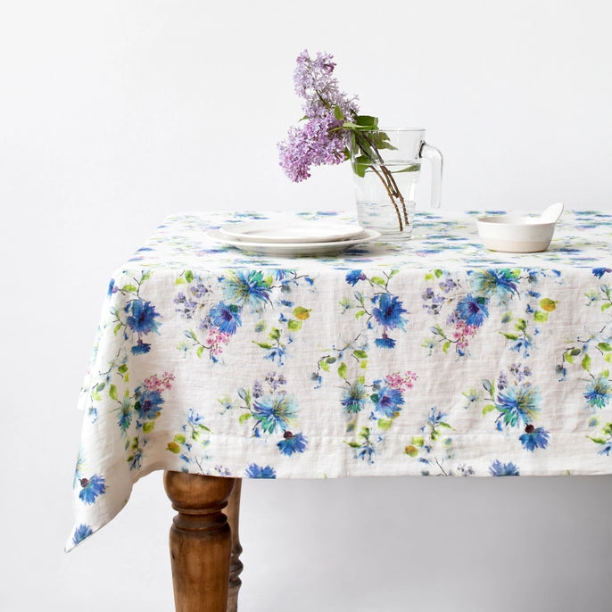 A tablecloth with a floral pattern, featuring various colors of flowers, placed on a wooden table with a vase of purple flowers and a small white plate on top of it, on a white background.