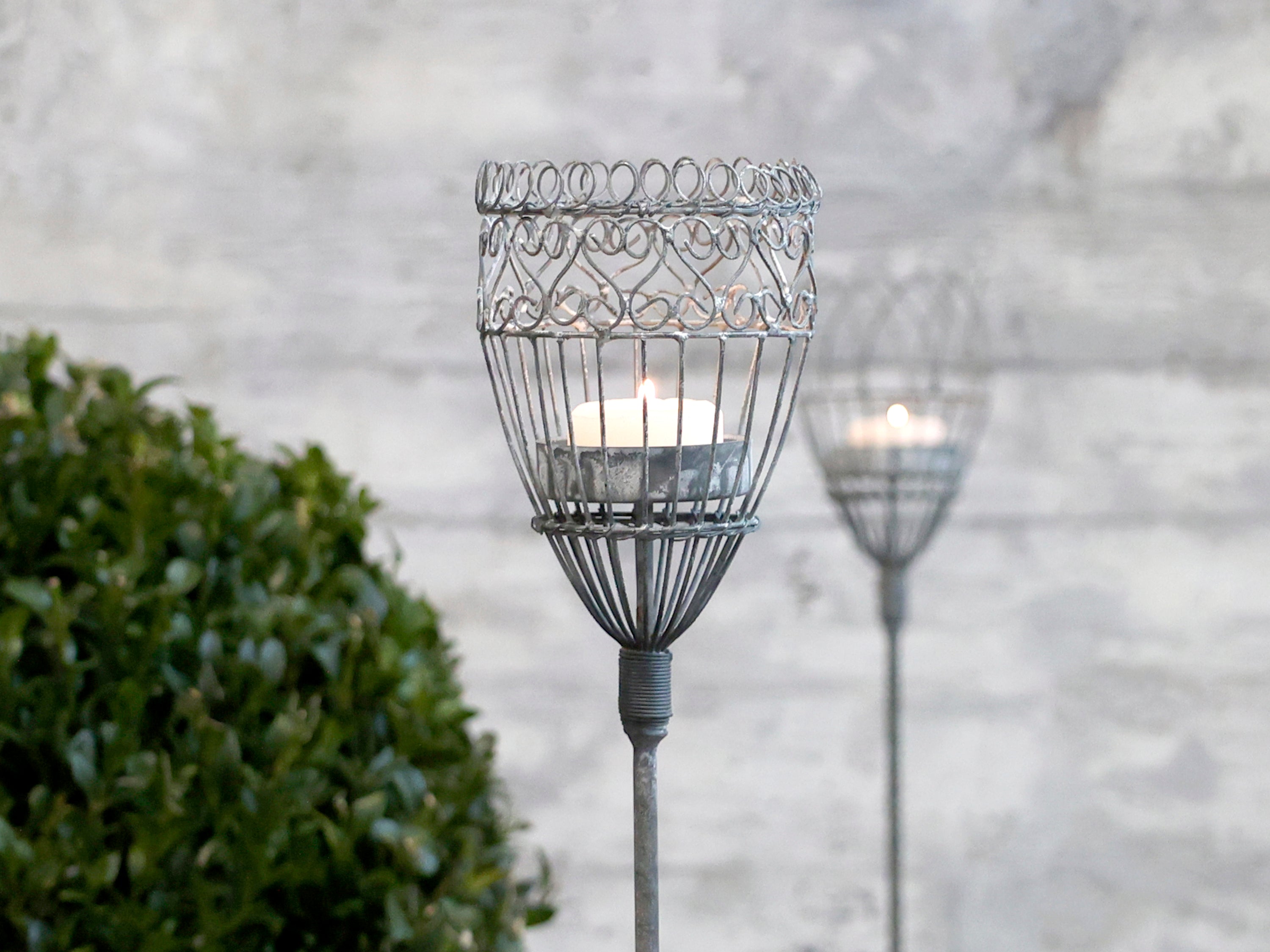 Close up of antique grey metal garden candle spear with another blurred in background with green plant and stone wall