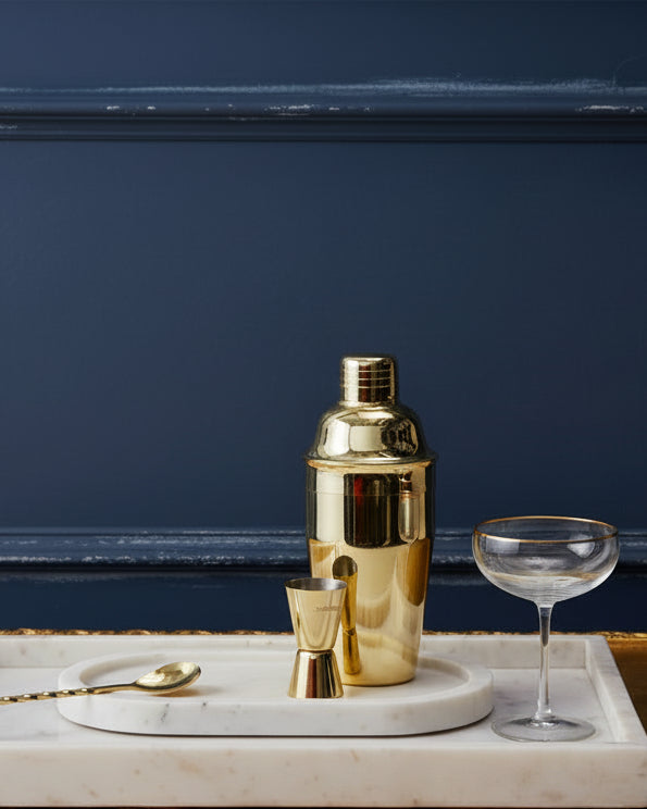 Gold-plated cocktail maker with jigger, gold spoon next to a gold-rimmed champagne coupe on a white marble tray against dark panelled wall