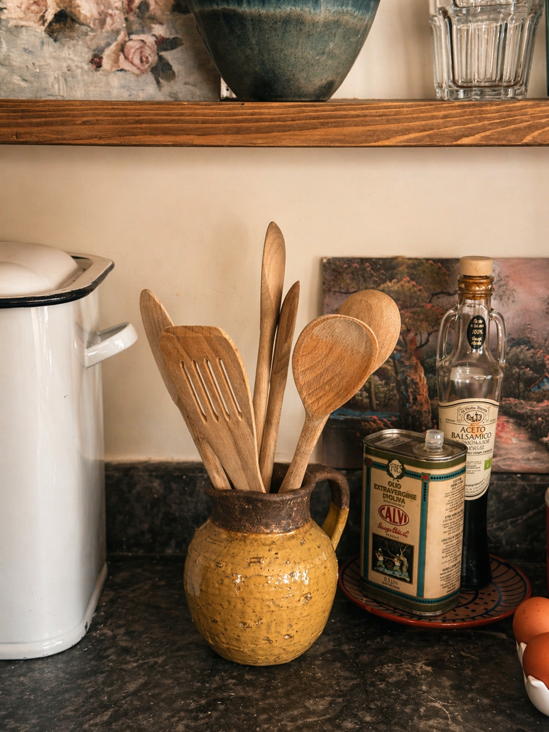 Wooden utensils in a yellow glazed terracotta jug on a kitchen counter next to bottles of olive oils, balsamic vinegar, eggs in a ceramic holder, and vintage white metal bread bin items in the background.